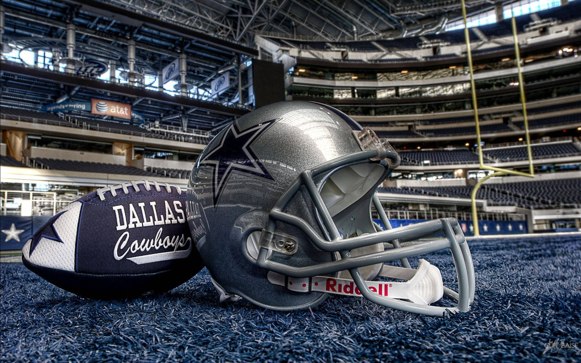 Dallas Cowboys Logo 桌布 壁紙 — 達拉斯牛仔隊的美式足球頭盔和球在明亮的燈光下，放置在空蕩蕩的體育館的藍色草地上。