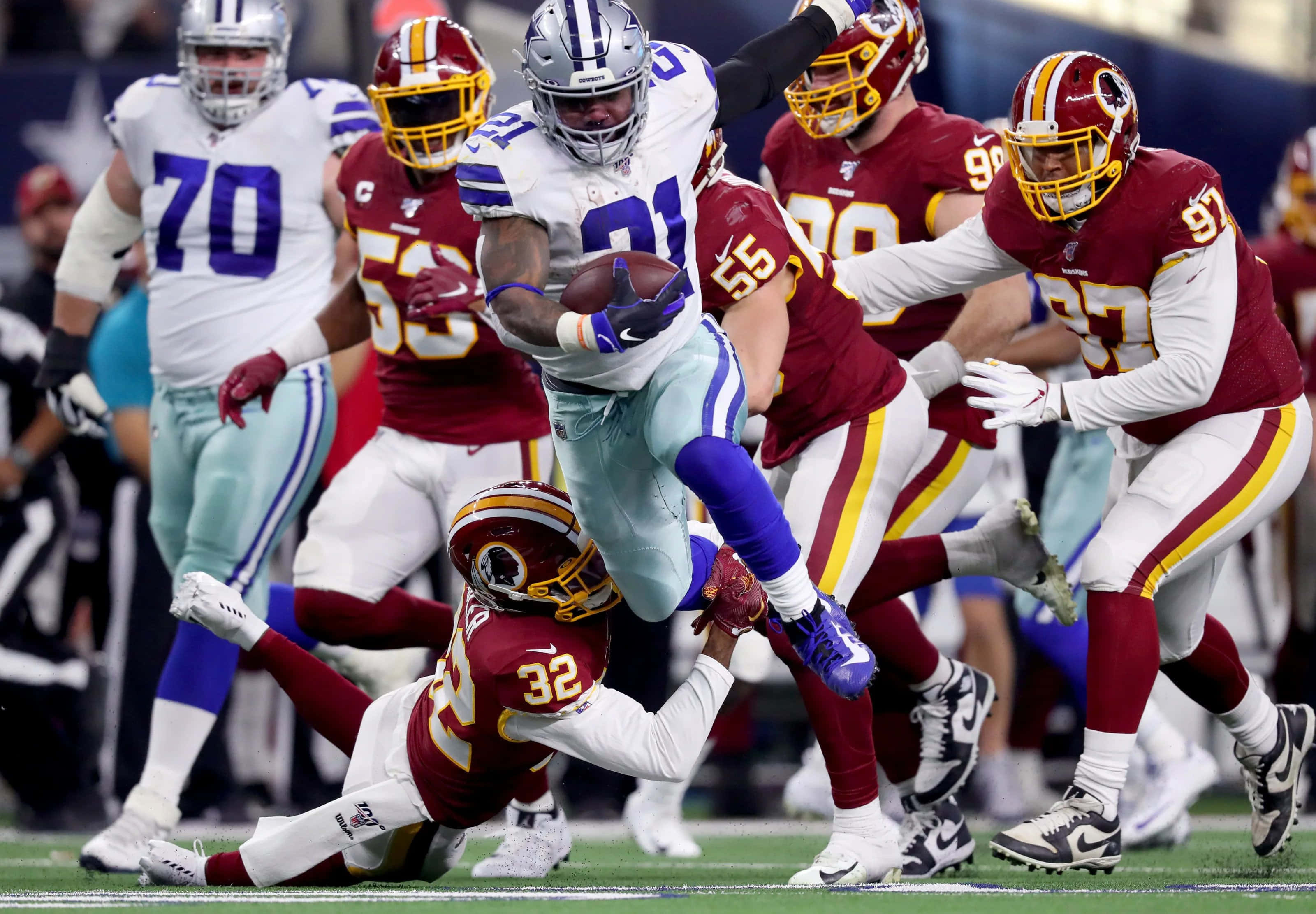 Washington Football 桌布 壁紙 — 達拉斯牛仔隊的跑鋒在NFL高風險比賽中緊抱橄欖球，躍過華盛頓美式足球隊的防守球員。
