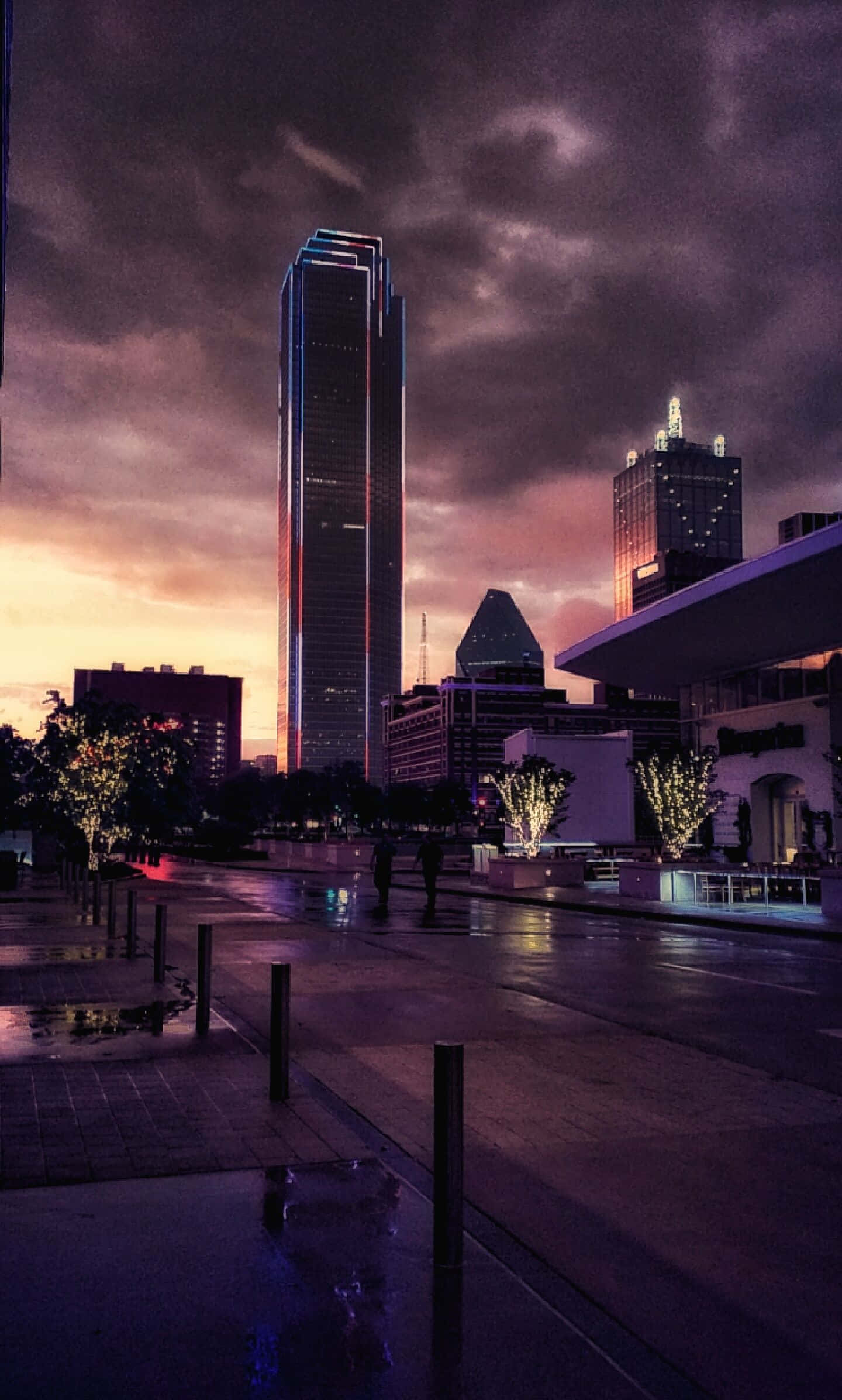 達拉斯縣背景與烏雲 壁紙 — 黃昏時分，濕漉漉的城市街道在戲劇性的紫色和橙色天空下，反射著高聳摩天大樓的霓虹燈光。