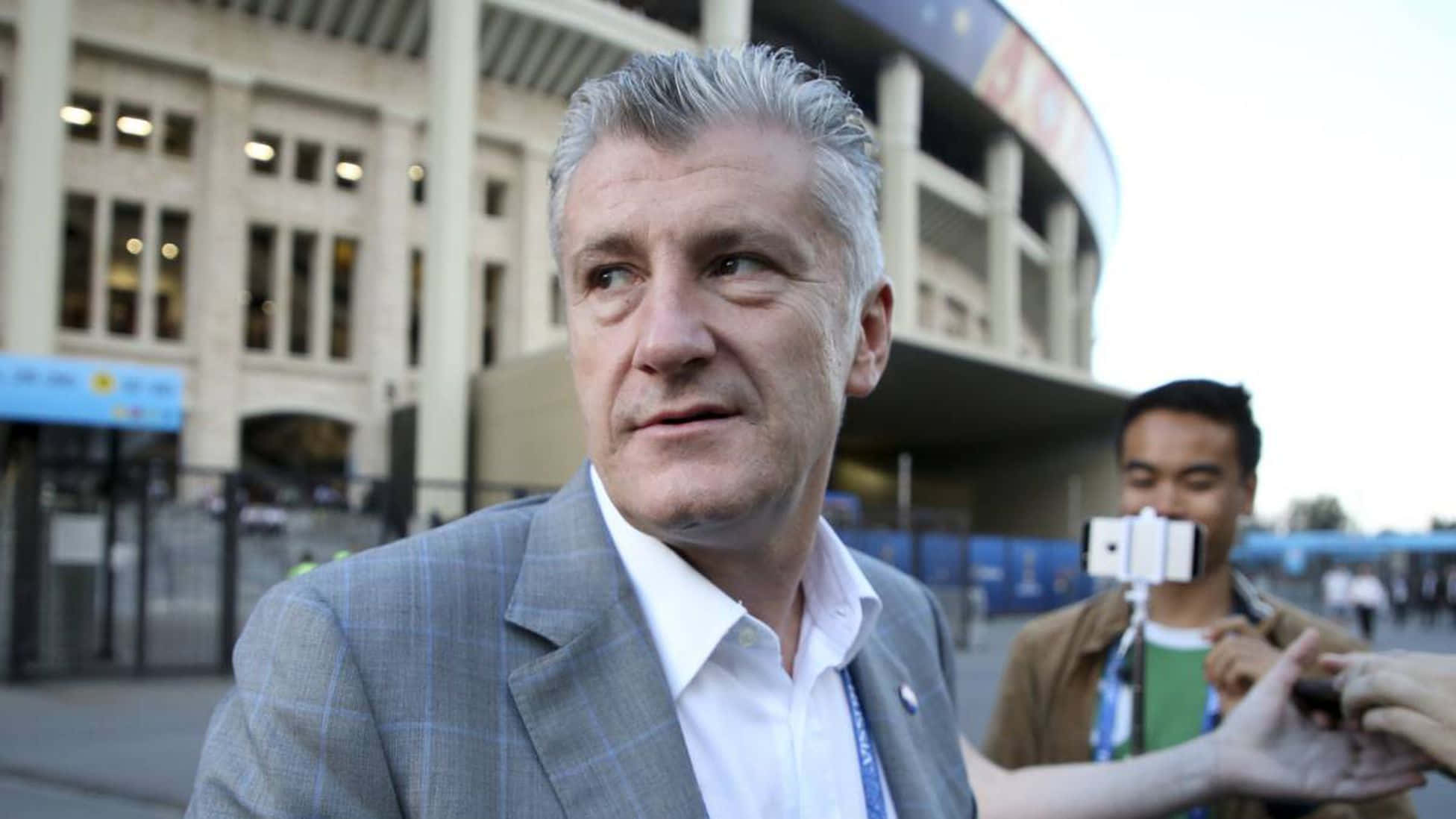 達沃·蘇克爾公開訪問即興畫面 壁紙 — 一名穿著灰色西裝的男子站在體育館外，一名粉絲舉起手機拍攝他。