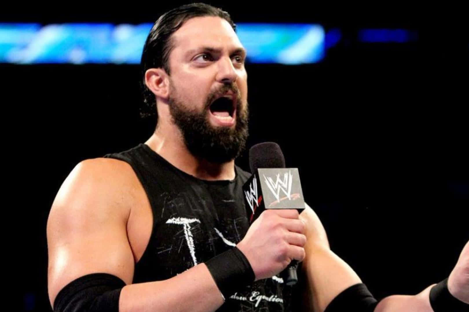 達米安·桑多對決The Miz 壁紙 — 一位肌肉發達、留鬍子的摔角手穿著黑色背心，在明亮的場館燈光下對著WWE麥克風大喊。