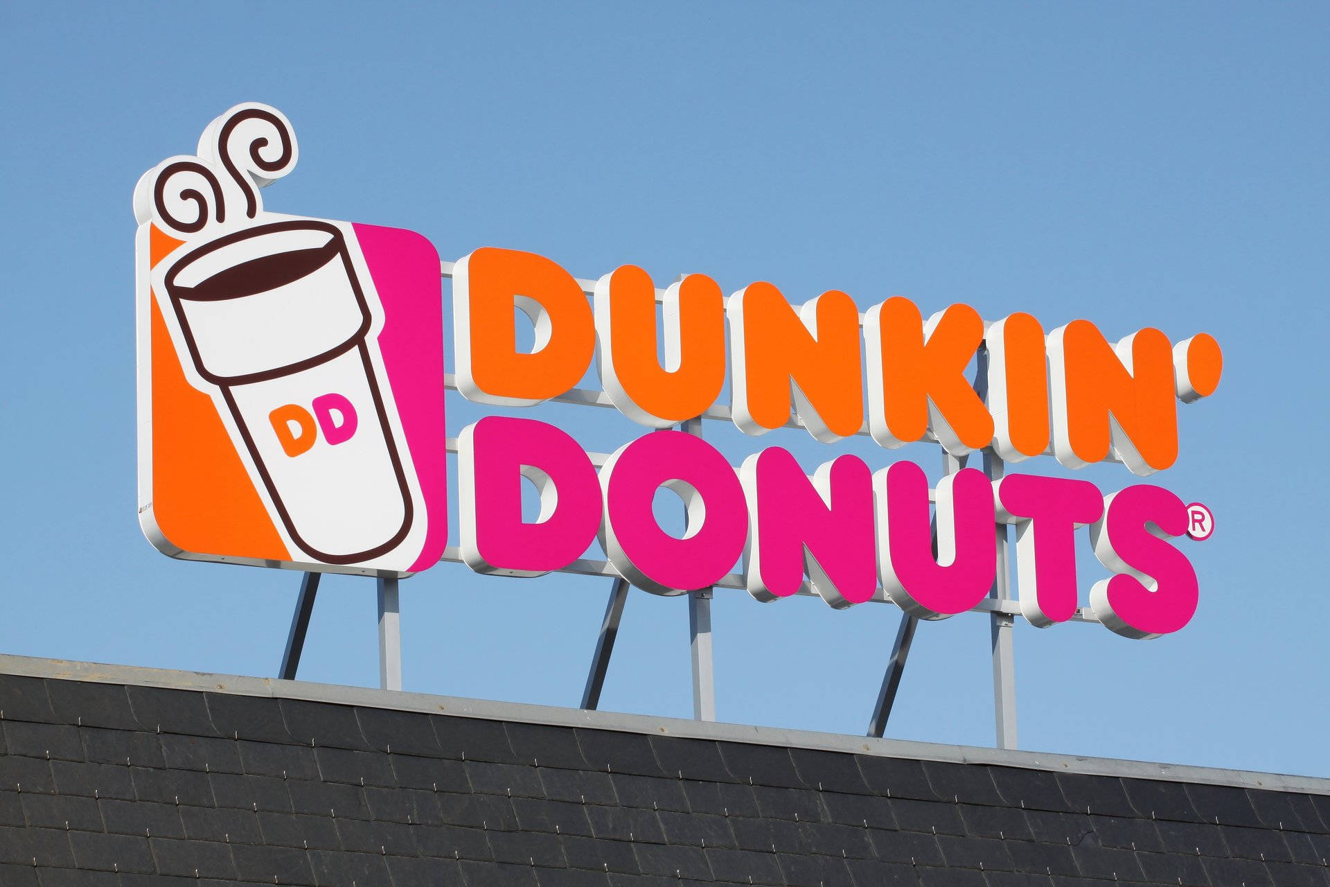 達美樂招牌 壁紙 — 一個明亮的Dunkin' Donuts招牌，帶有橙色和粉紅色的字樣以及咖啡杯標誌，在深色屋頂上方清澈的藍天下顯得格外醒目。