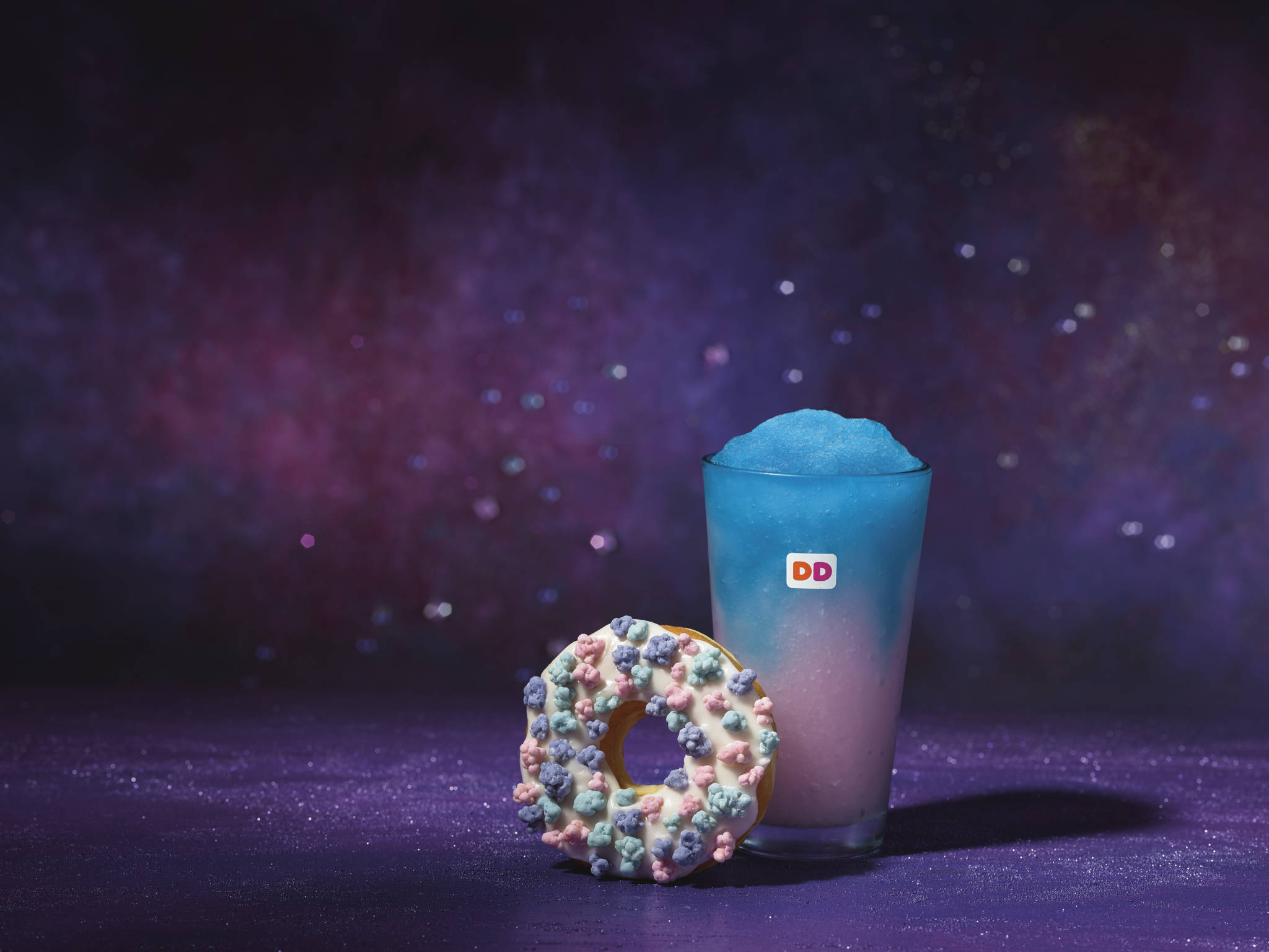 達美樂星際主題 壁紙 — 一杯藍色思樂冰飲品和一個灑上糖粒的冬甩，放在閃閃發光的紫色表面上，背景是宇宙星空。