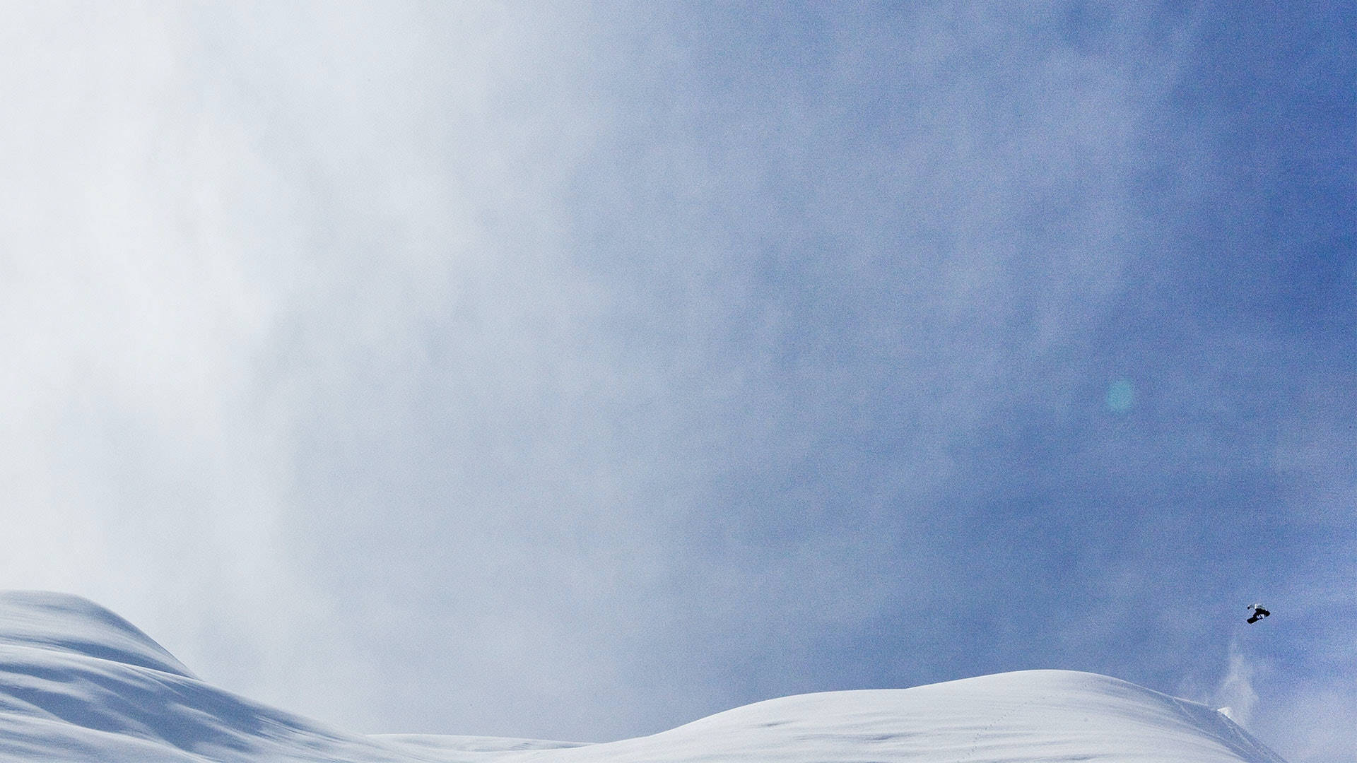 遠眺滑雪者 壁紙 — 一名滑雪板運動員在晴朗的藍天下，飛越廣闊的雪山，穿梭於清爽的冬日空氣中。