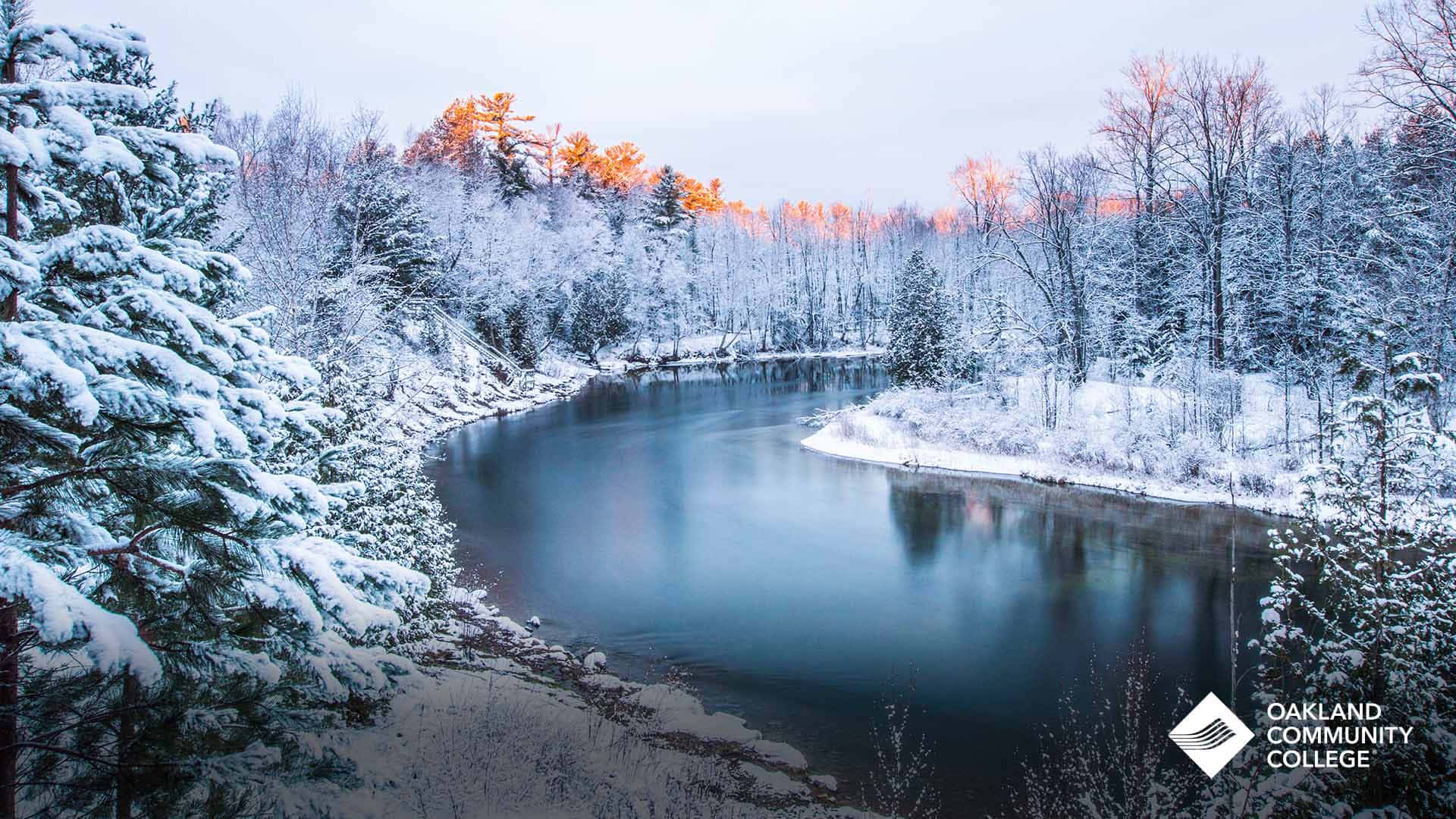 遠眺雪覆山巒，冬日仙境美景 壁紙 — 一條寧靜的冬季河流蜿蜒穿過白雪覆蓋的松樹和白樺樹，金色的日落光芒透過森林樹冠閃耀。