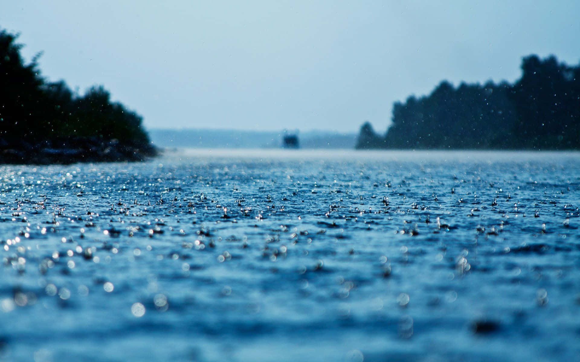 遠處群山環繞的湖景 壁紙 — 雨滴在廣闊的藍色湖面上泛起漣漪，天空朦朧，遠處有樹木成排的湖岸。