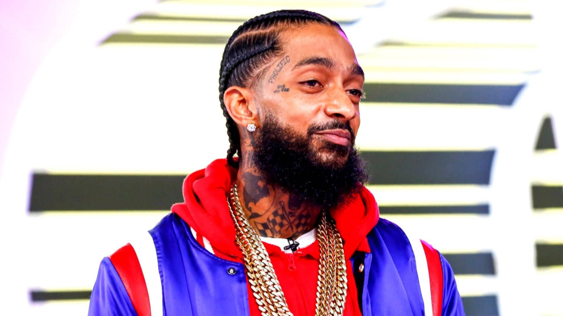 遠見卓識的饒舌歌手 Nipsey Hussle 壁紙 — 一個有辮子頭髮和紋身的男人，在條紋背景前穿著藍色外套下的紅色連帽衫和金鏈。
