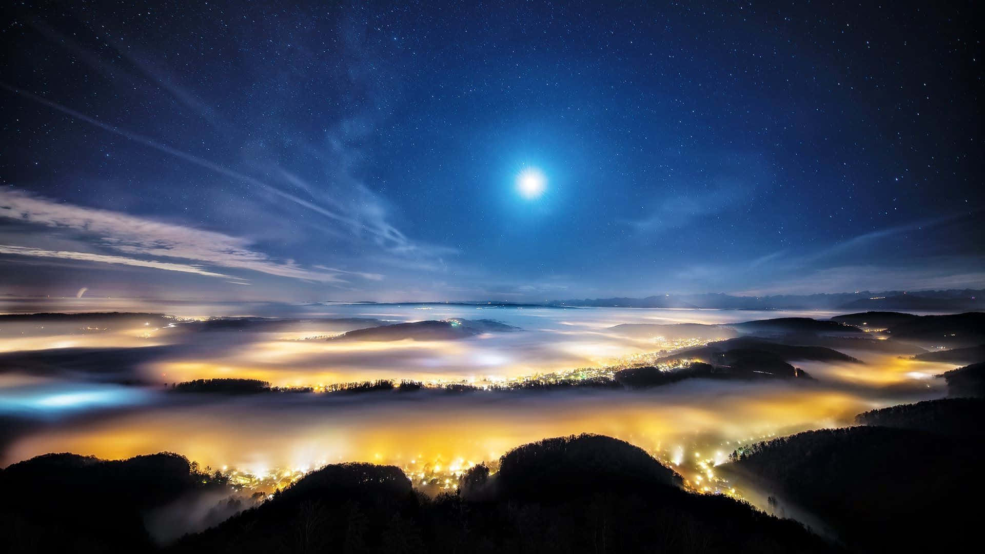 遠離白日喧囂，探索夜的寧靜 壁紙 — 一輪明亮的月亮照耀在黑暗森林山丘之間的發光雲海之上，下方是星空夜空。