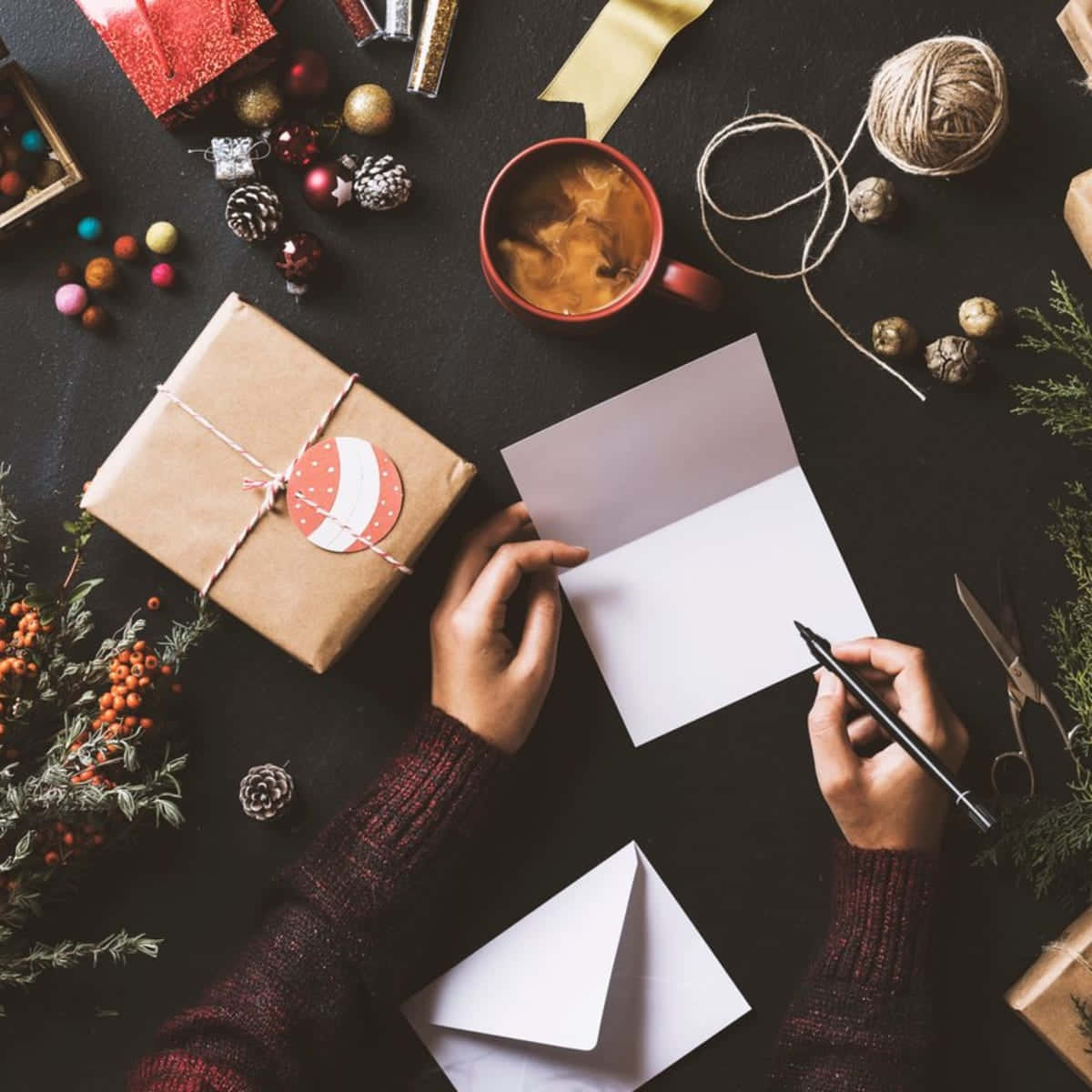 適合寄送溫暖祝福的雪景聖誕卡 壁紙 — Overhead view of hands writing a holiday card beside a wrapped gift and festive decorations on a dark table.
