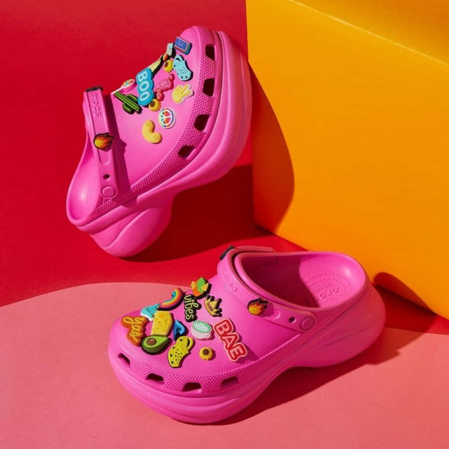 在色彩繽紛的幾何背景上，裝飾著有趣飾物的粉紅色Crocs。