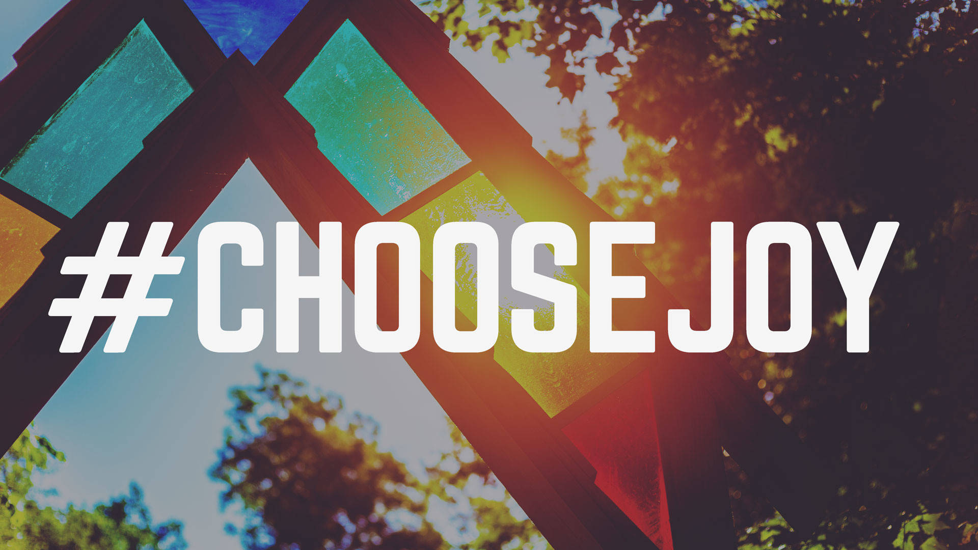選擇歡樂標籤 壁紙 — 粗體白色文字 hashtag choosejoy 覆蓋在充滿活力的彩繪玻璃結構上，背景有陽光照射的樹木。