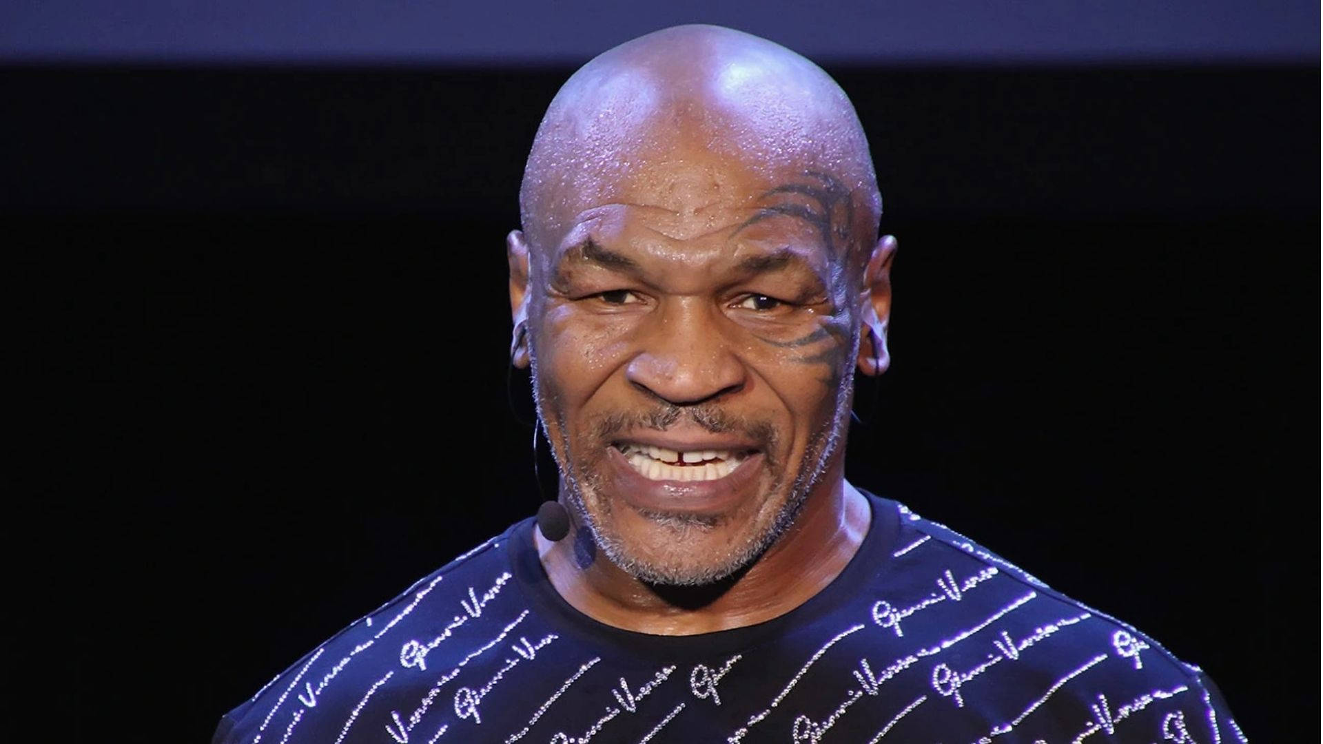 邁克·泰森 談話 壁紙 — Mike Tyson 在舞台上燦爛地笑著，穿著 Gucci 襯衫，在聚光燈下，背景為黑色。