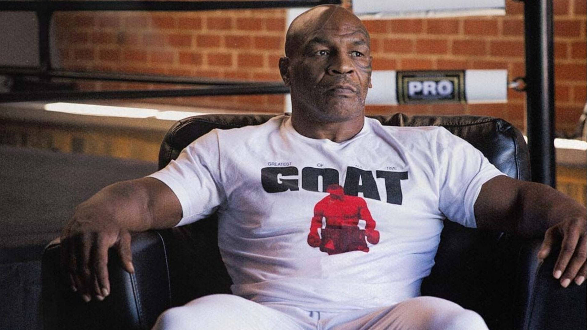 邁克·泰森坐在沙發上 壁紙 — Mike Tyson 自信地坐在黑色皮椅上，穿著一件印有 'GOAT' 和紅色拳擊人物圖案的白色 T 恤。