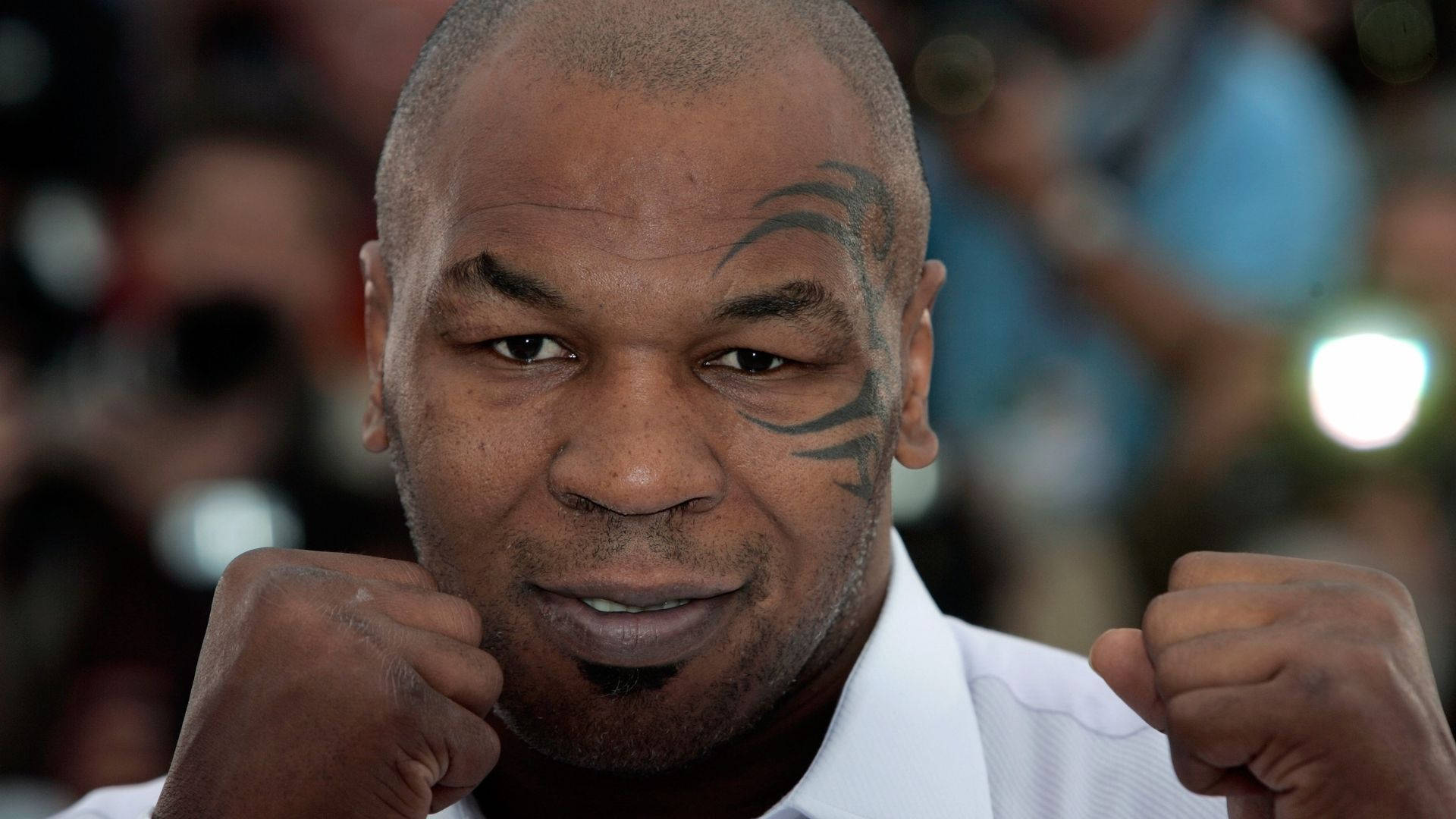 邁克·泰森招牌拳擊姿勢 壁紙 — Mike Tyson 穿著白色襯衫，在模糊的人群背景下，舉起拳頭，凝視著。