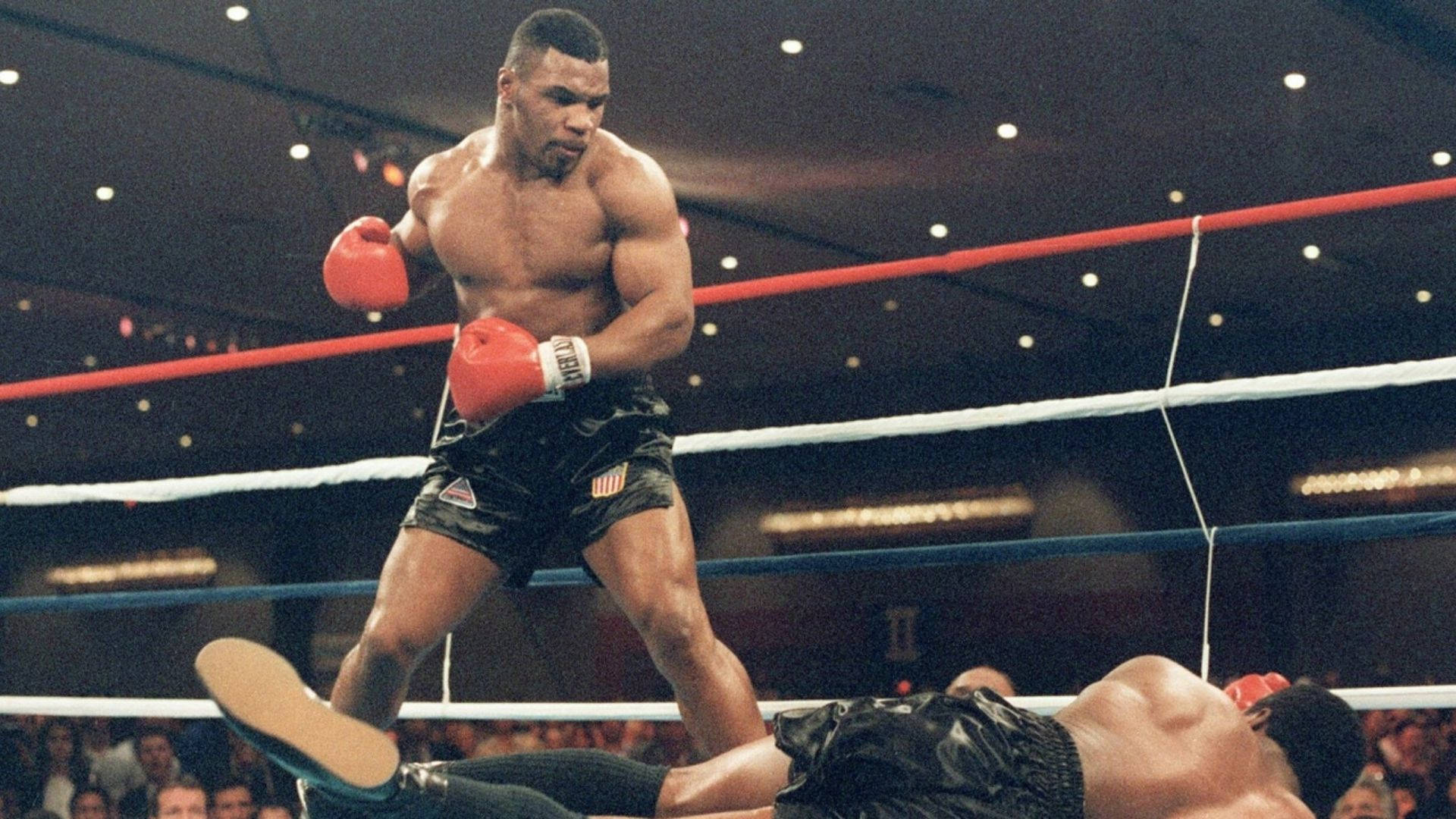 邁克·泰森擊倒對手 壁紙 — Mike Tyson 在擂台上勝利地站著，而他的對手在明亮的場館燈光下被打敗躺在地上。