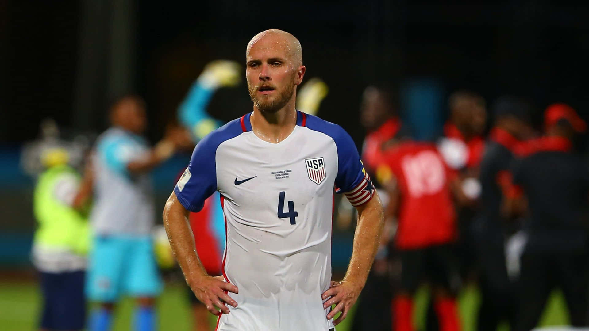 邁克爾·布拉德利在FIFA世界盃資格賽 壁紙 — 一位光頭的美國足球員身穿白色球衣，號碼4，雙手叉腰站在夜間球場上。