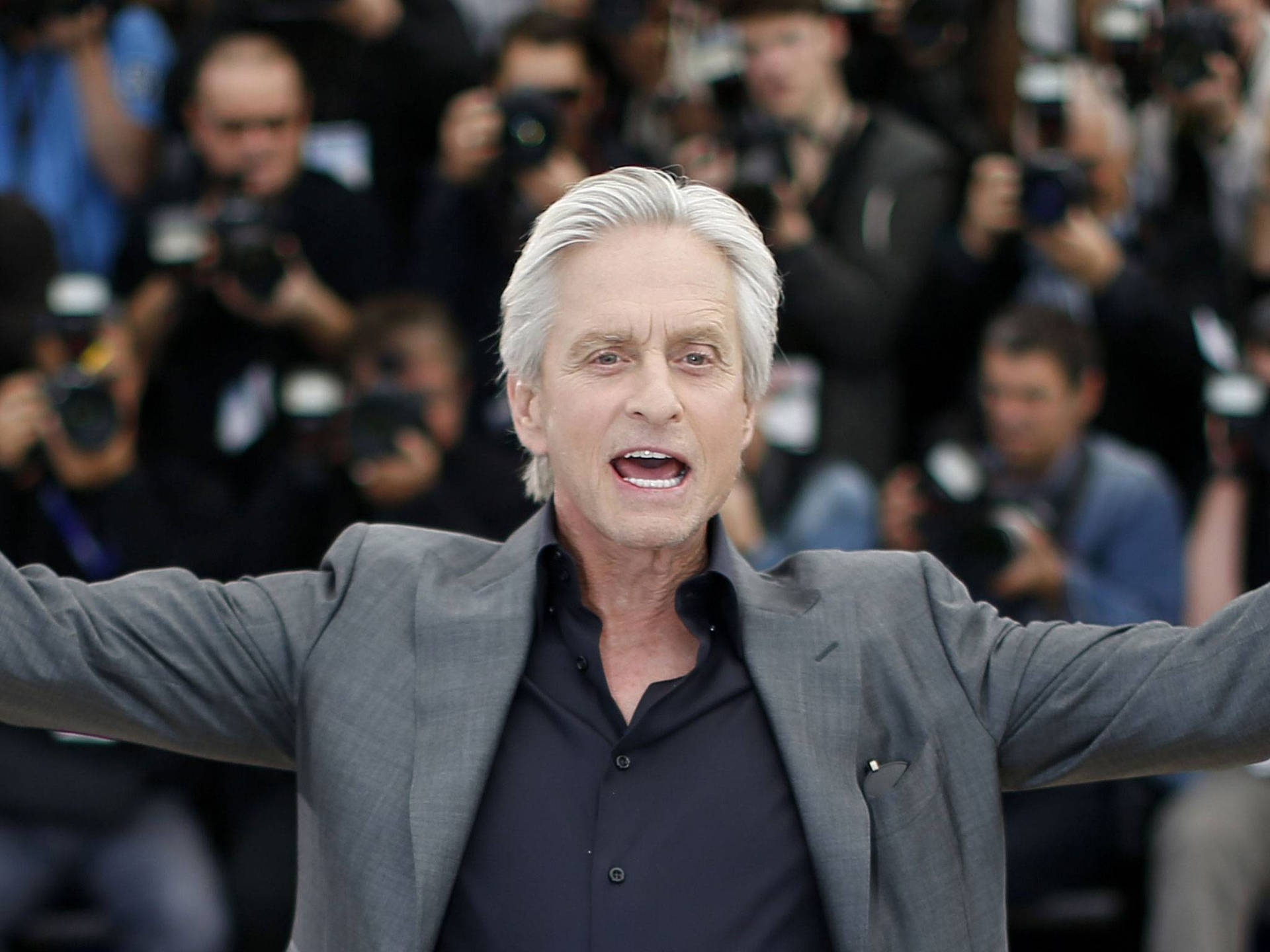 身穿灰色西裝的男士在公開活動中伸展雙臂，身後有攝影師。