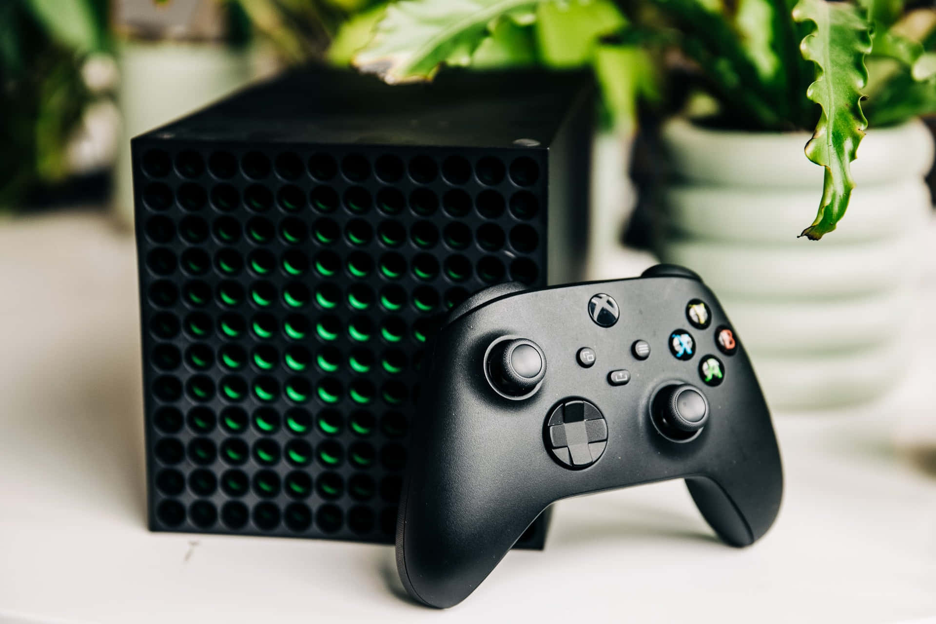 黑色 Xbox 主機和控制器，帶綠色燈光，旁邊是白色桌子上的室內植物。