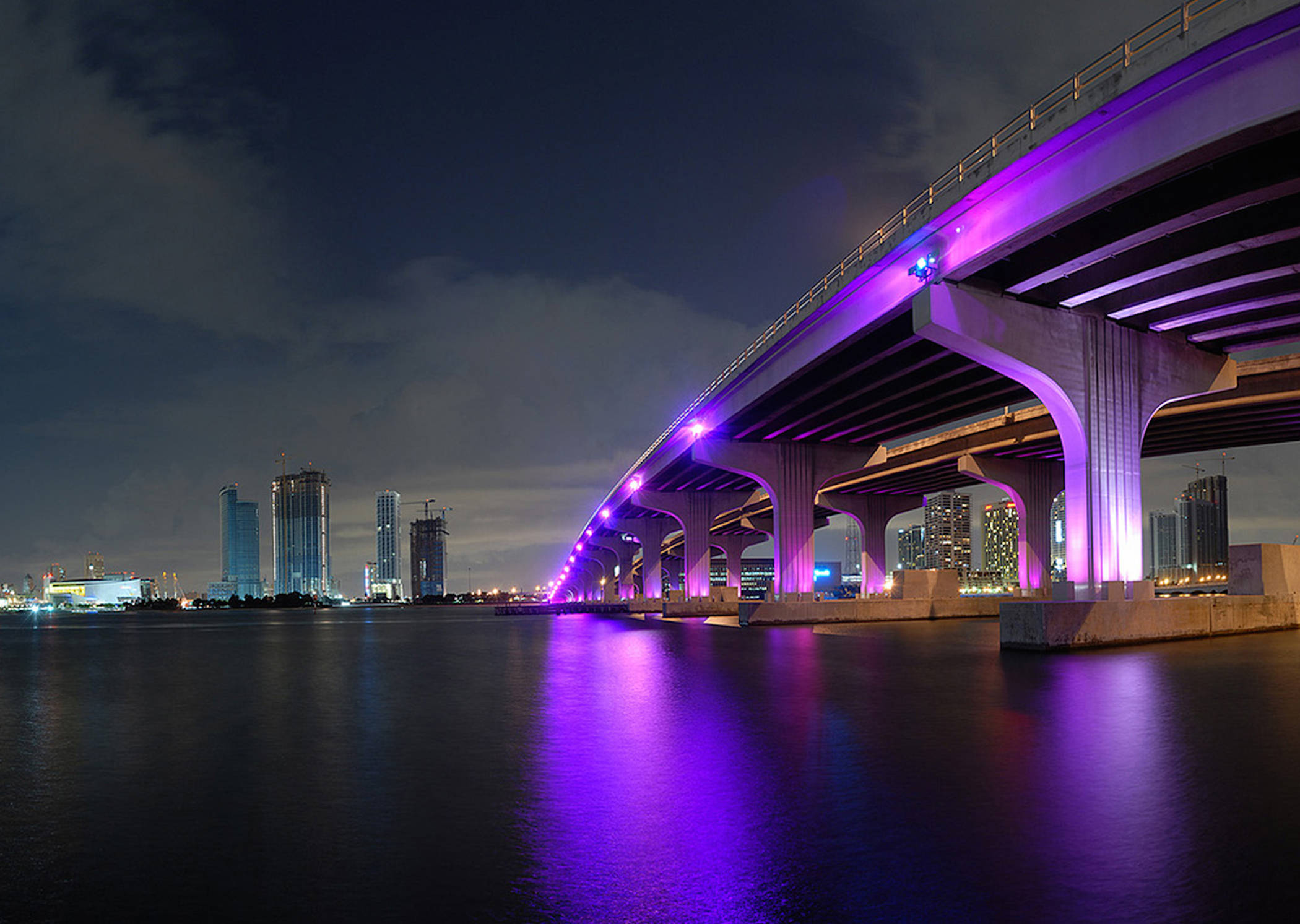 邁阿密橋樑紫光夜景 壁紙 — 一座宏偉的照明橋樑沐浴在紫色燈光中，橫跨平靜的水面，倒映著黑暗夜空下的城市摩天大樓。