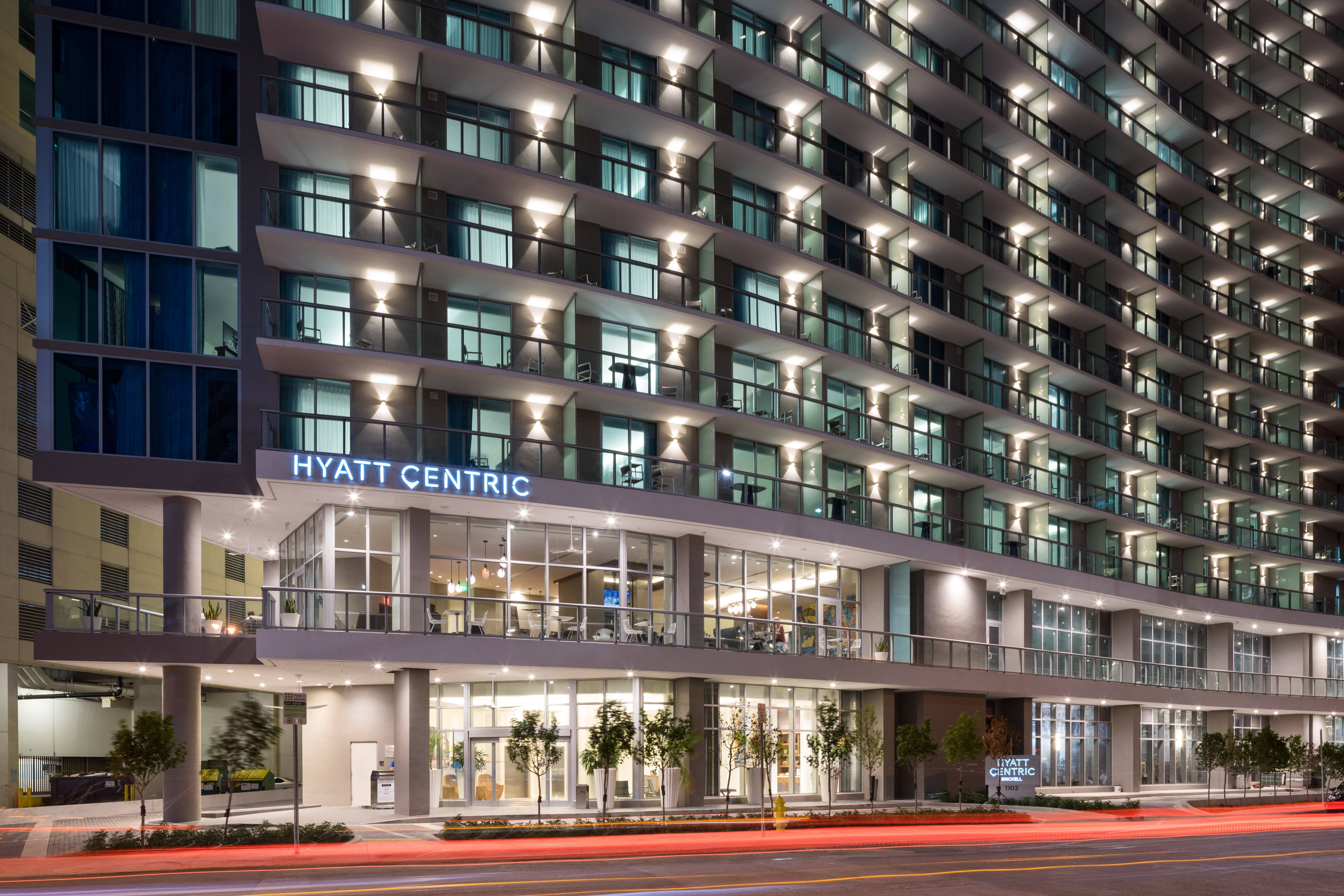 邁阿密海灘的海雅特中心酒店 壁紙 — 一座現代化的Hyatt Centric酒店建築在夜晚亮起，窗戶發光，下方有紅色交通燈光的痕跡。