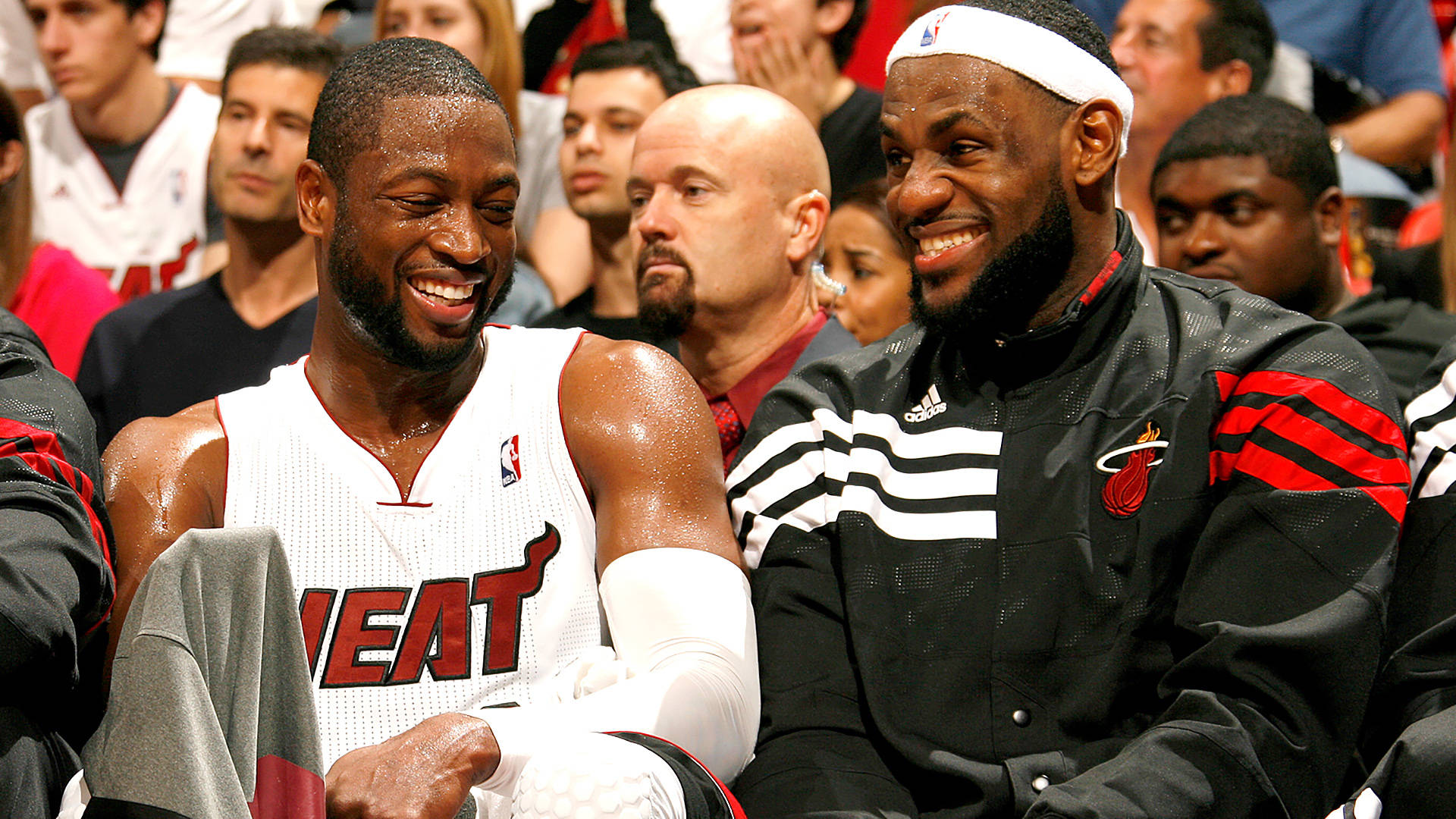 邁阿密熱火隊 LeBron 與 Wade 壁紙 — 兩位微笑的邁阿密熱火籃球運動員在比賽期間坐在後備席上，背景有球迷，分享歡樂時刻。