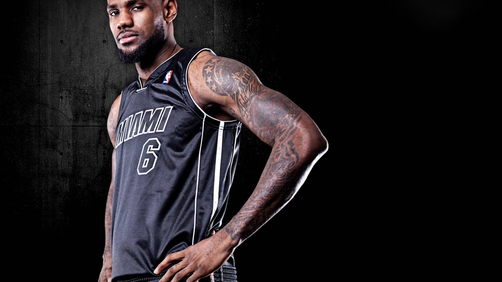 邁阿密熱火隊 LeBron 黑色球衣 壁紙 — 一位肌肉發達的籃球運動員身穿邁阿密熱火隊球衣，自信地站在深色紋理牆壁前，手臂上的紋身清晰可見。