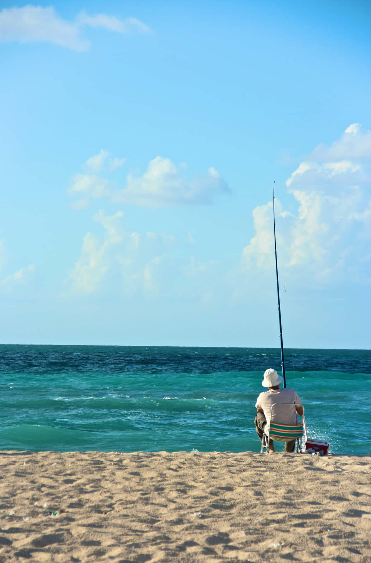 一位年長男子在陽光明媚的海灘上獨自釣魚，海水碧綠，天空湛藍。