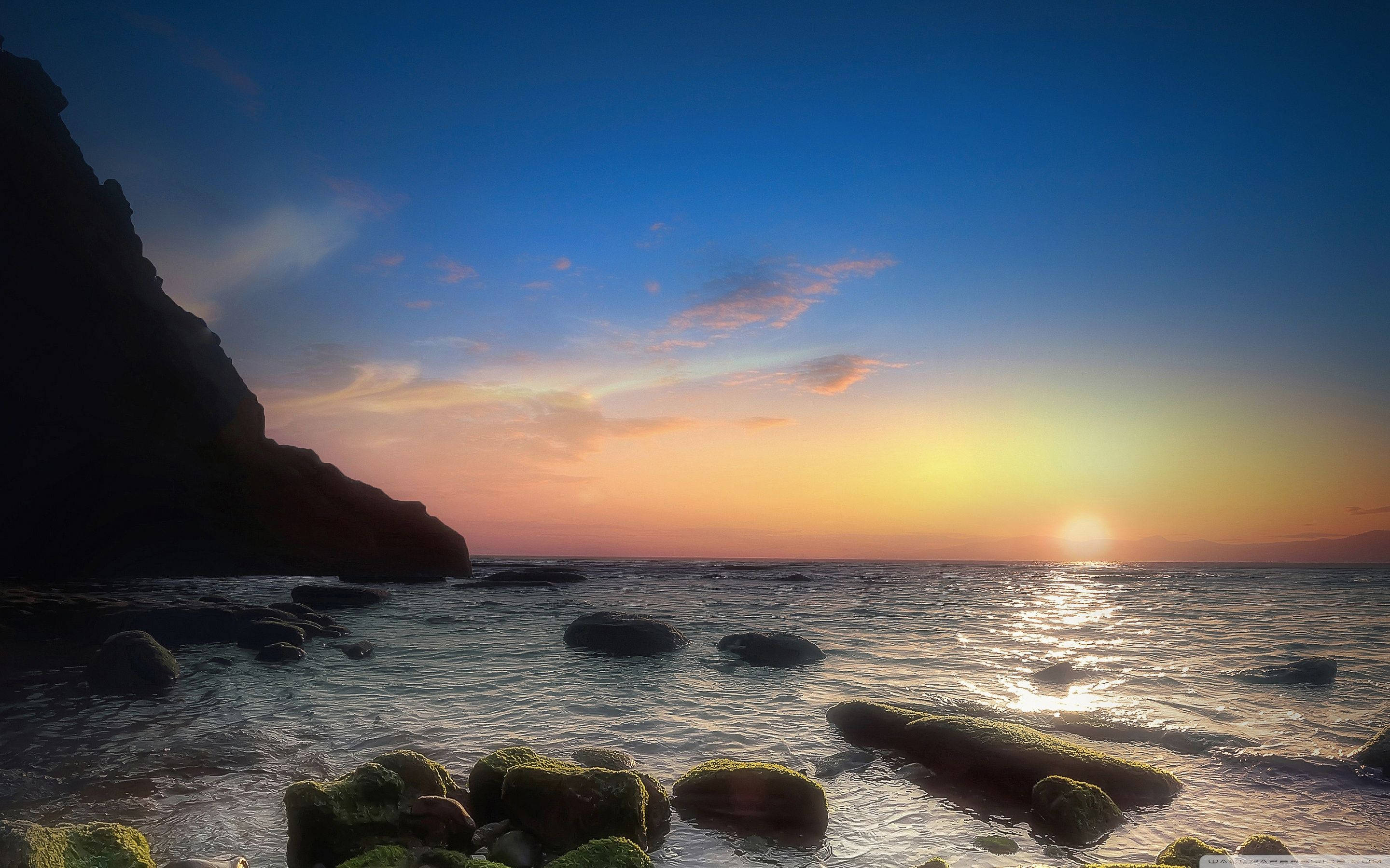 那不勒斯海景 壁紙 — 夕陽以溫暖色調塗抹天空，海浪輕輕拍打著黑暗懸崖旁長滿苔蘚的岩石。