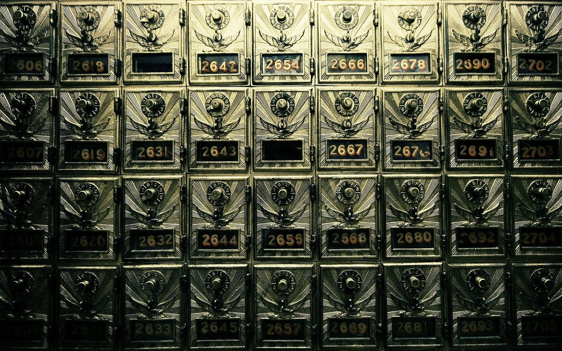 郵政局儲物櫃保險庫 壁紙 — 暖色燈光下，復古黃銅郵箱的特寫，帶有翼形徽章和編號槽，以網格排列。