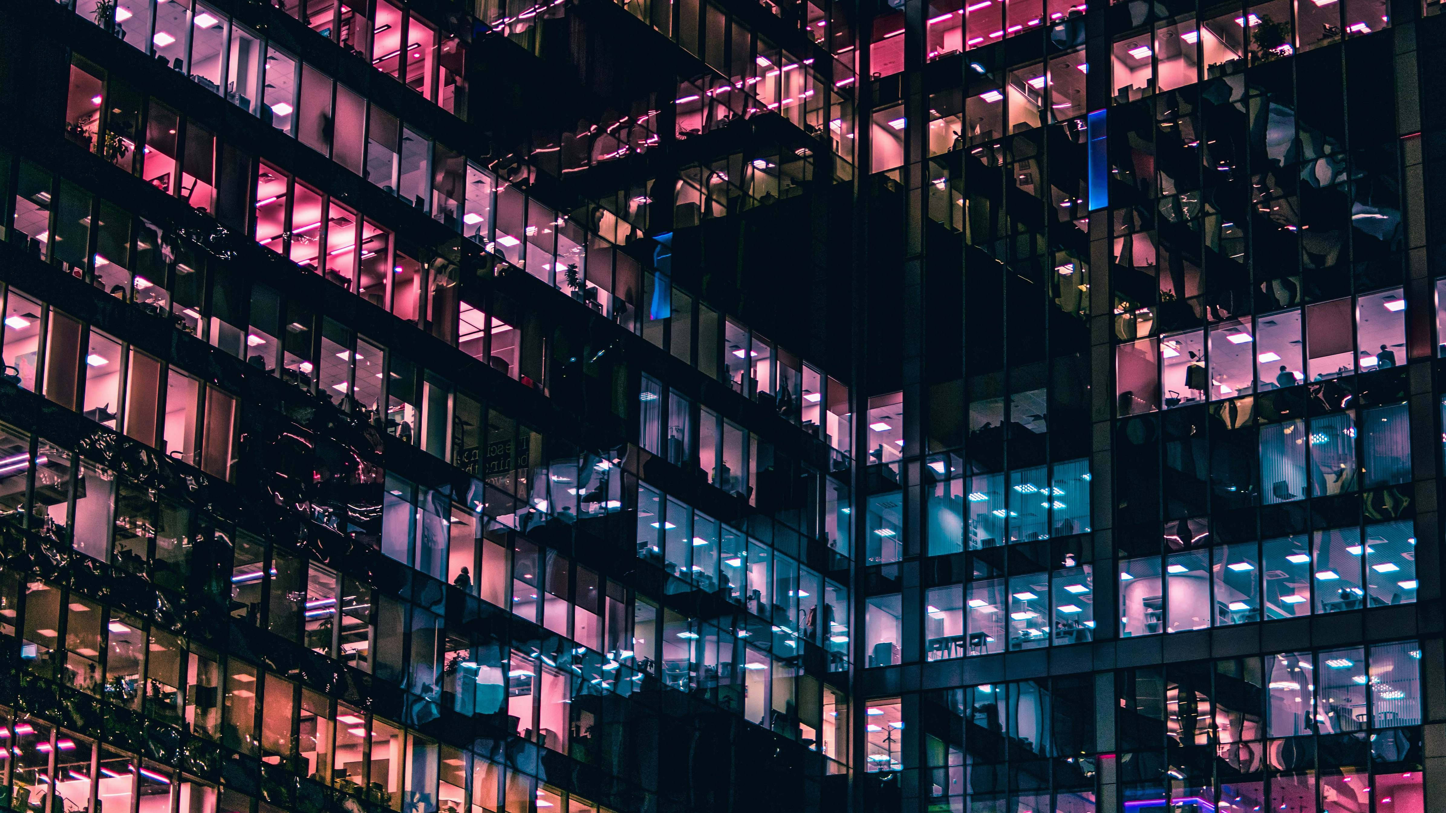 都市復古莫斯科俄羅斯 壁紙 — 夜晚一座高聳的玻璃摩天大樓，帶有發光的粉紅色和藍色辦公室燈光，顯露出內部的工人。