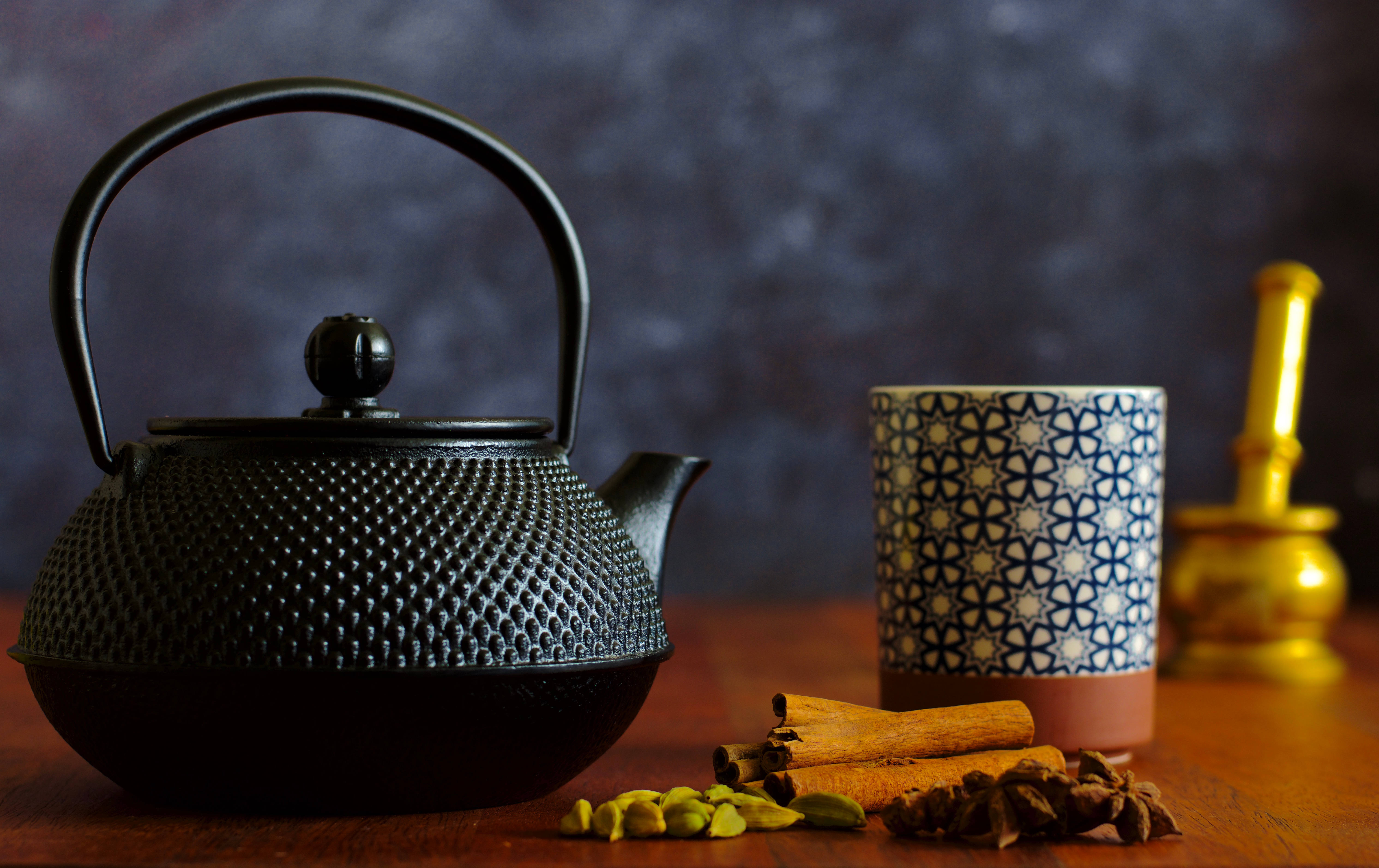 鄉村秋日茶香 壁紙 — 一個黑色紋理茶壺旁邊放著一個圖案杯子，杯中有肉桂棒和香料，放在木桌上，背景是深色紋理。