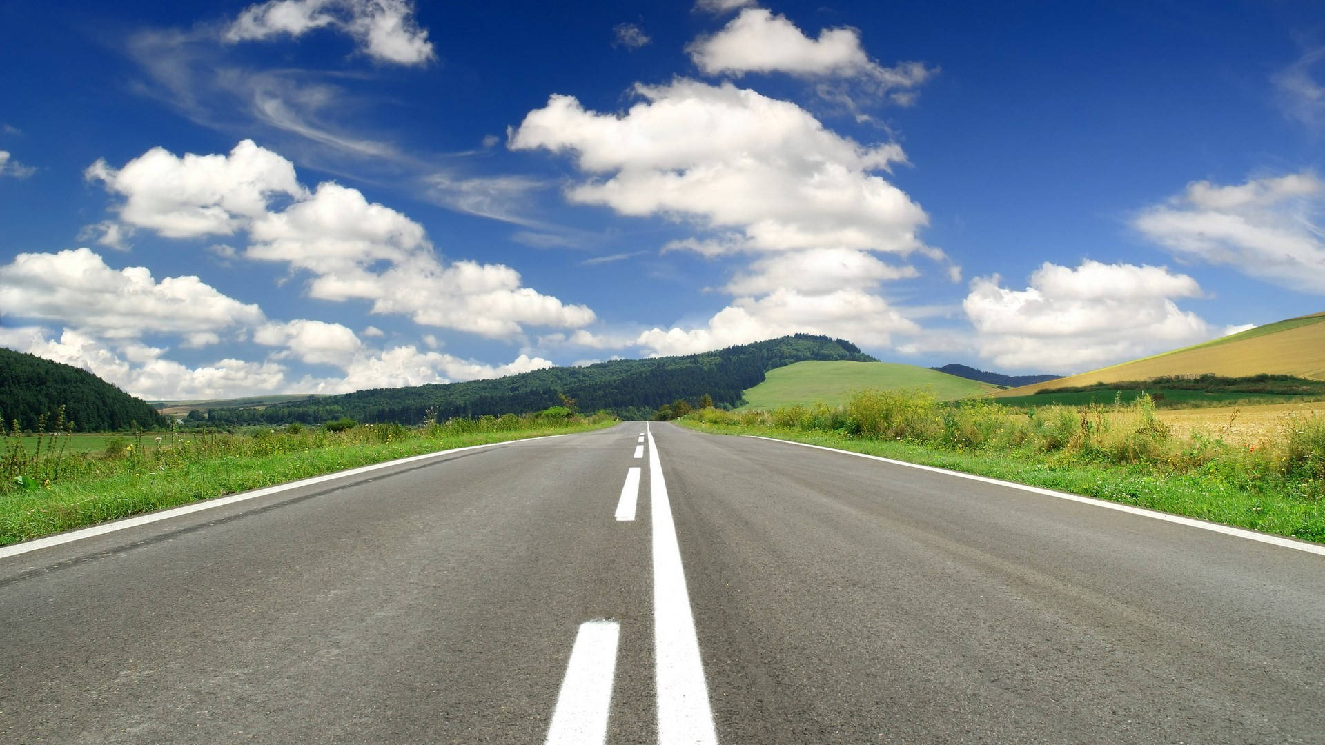 鄉村道路風景照 壁紙 — 一條筆直的瀝青道路在晴朗藍天和蓬鬆白雲下，伸向遠方的綠色山丘。