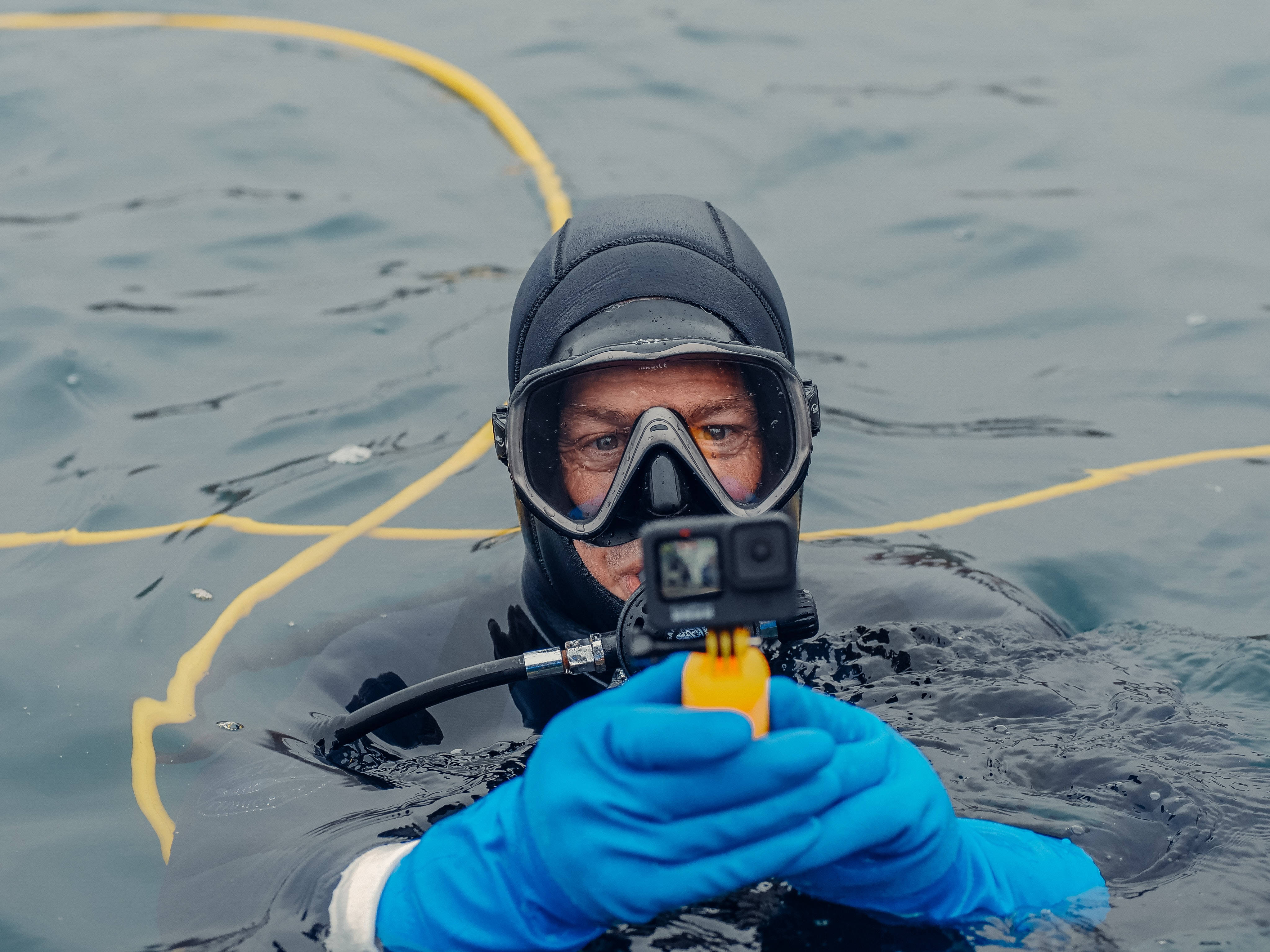 配GoPro的潛水員 壁紙 — 一名身穿黑色潛水衣和藍色手套的潛水員，手持GoPro相機在水面上方，身後拖著一條黃色繩索。