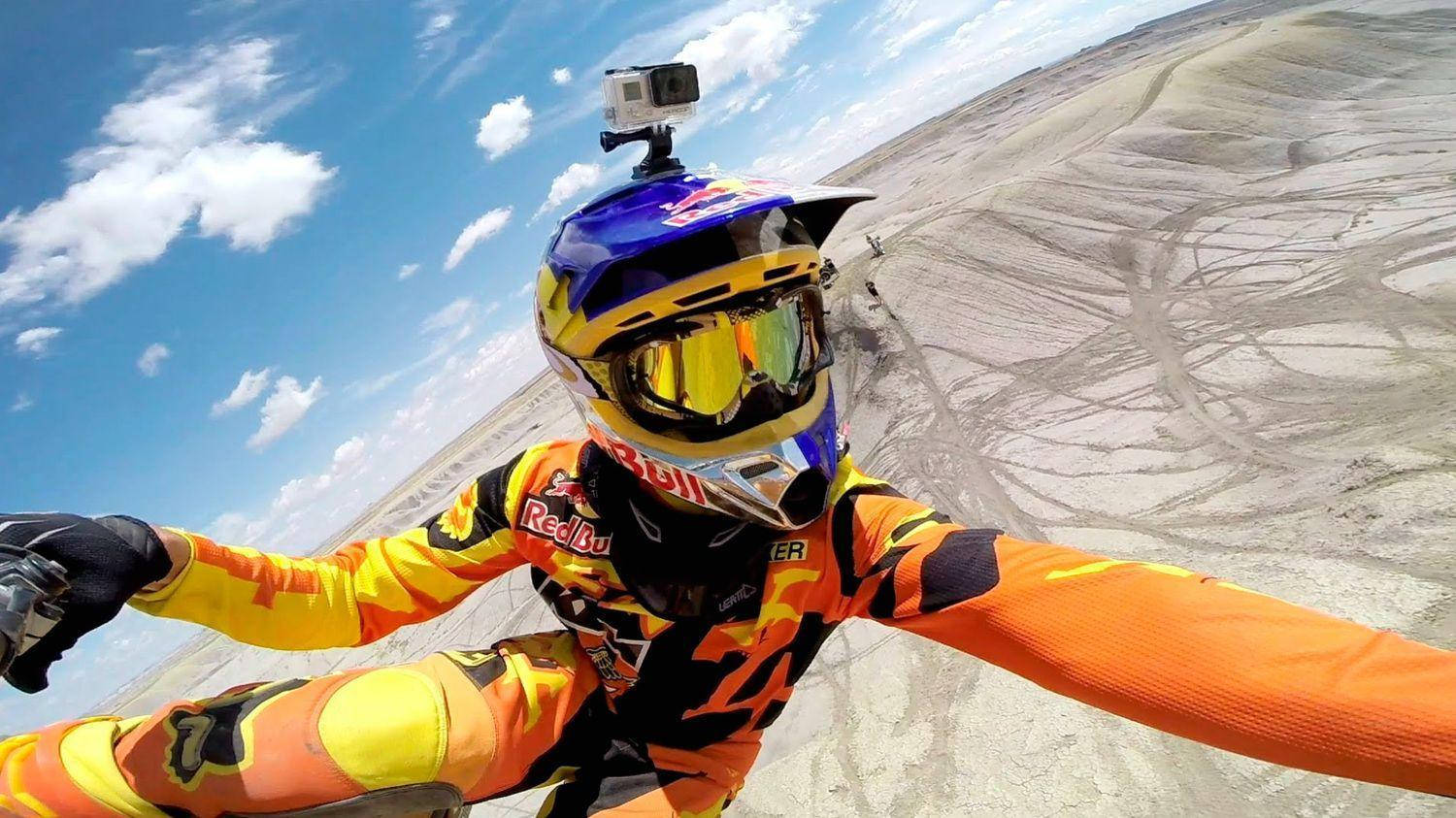配備 GoPro 的跳傘員頭盔 壁紙 — 一名身穿鮮豔裝備的越野電單車手，在塵土飛揚的賽道上，於藍天白雲下自拍。