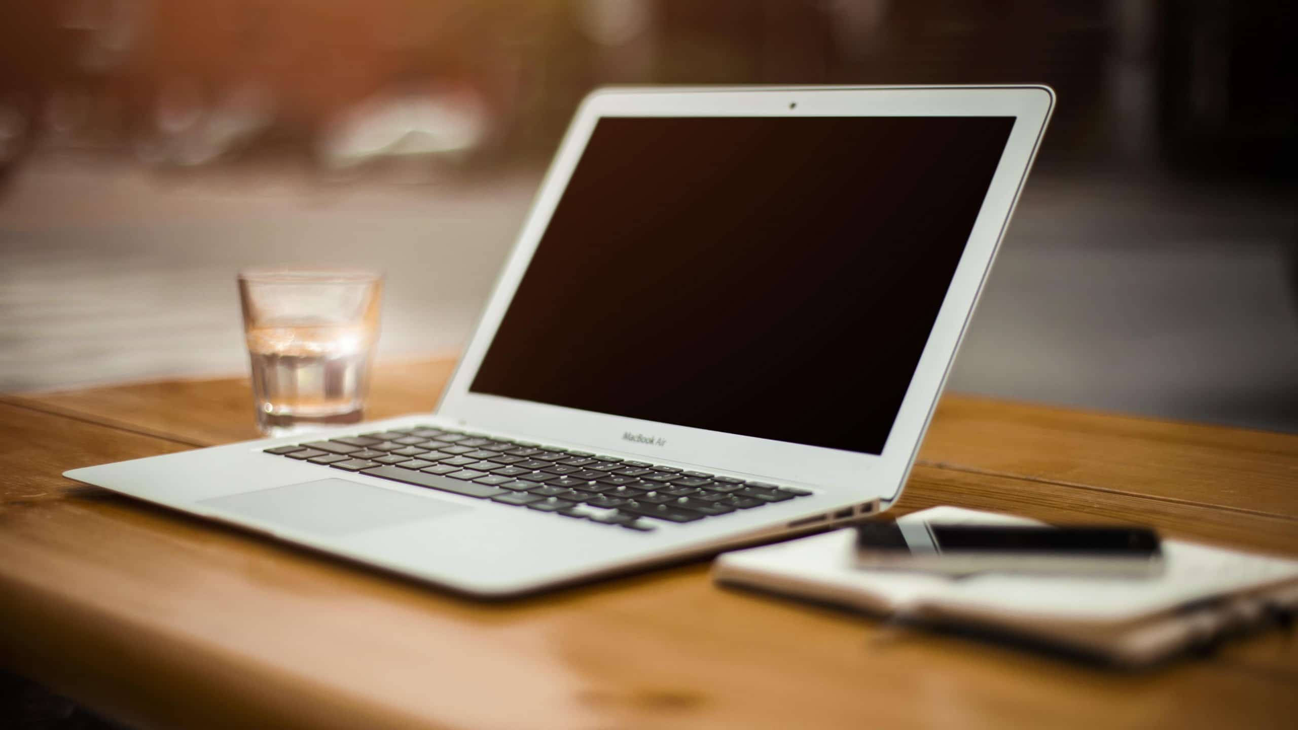 配備MacBook Air的辦公桌 壁紙 — 一部銀色手提電腦打開放在木桌上，旁邊有一杯水和一本附有筆的筆記簿。