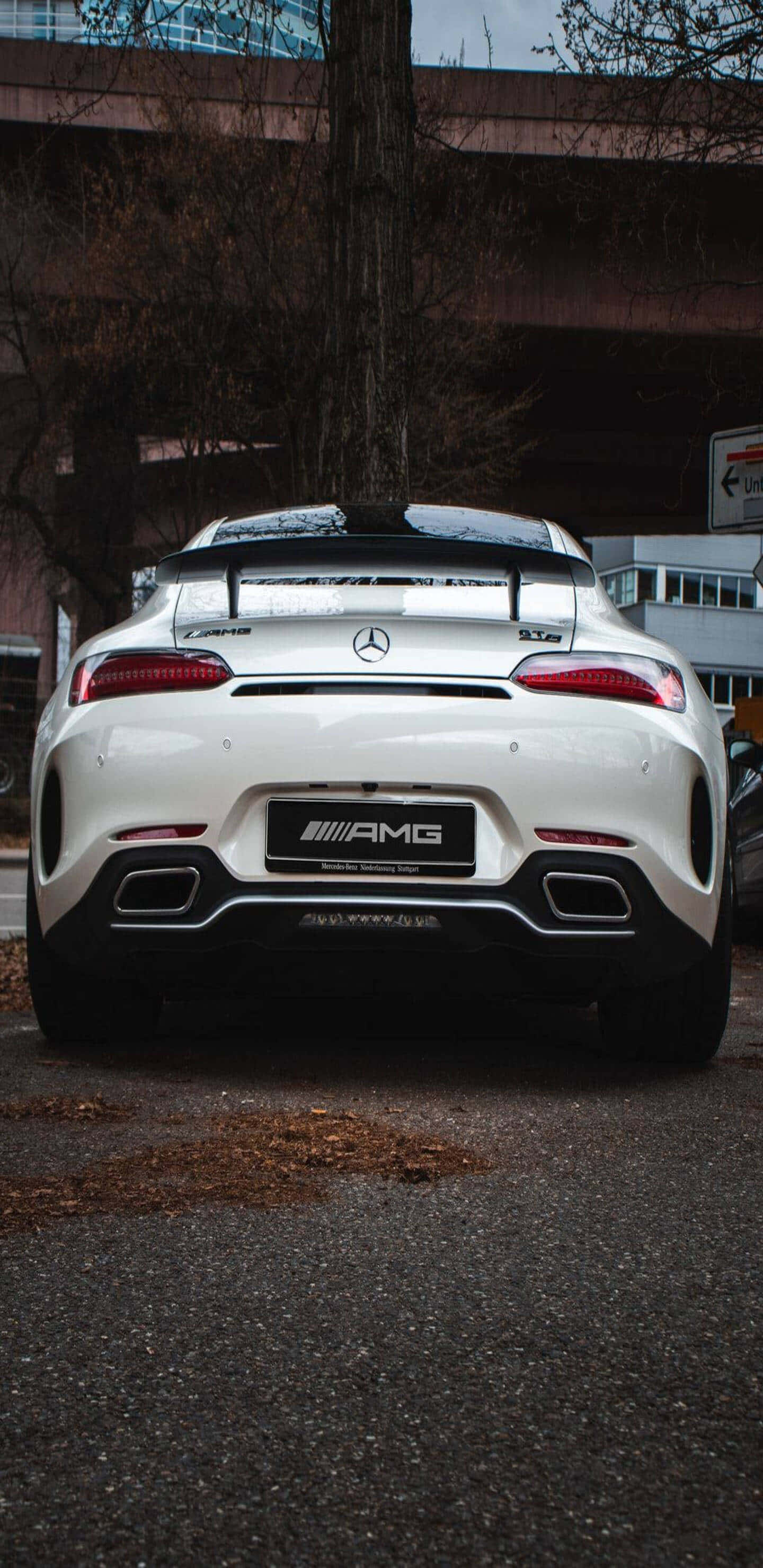 配備黑色尾翼的白色梅賽德斯-奔馳 Pixel 3XL AMG GT-R背景 壁紙 — 從後方觀看一輛白色Mercedes-AMG GT R跑車停在天橋下，背景有光禿的樹木和城市建築。