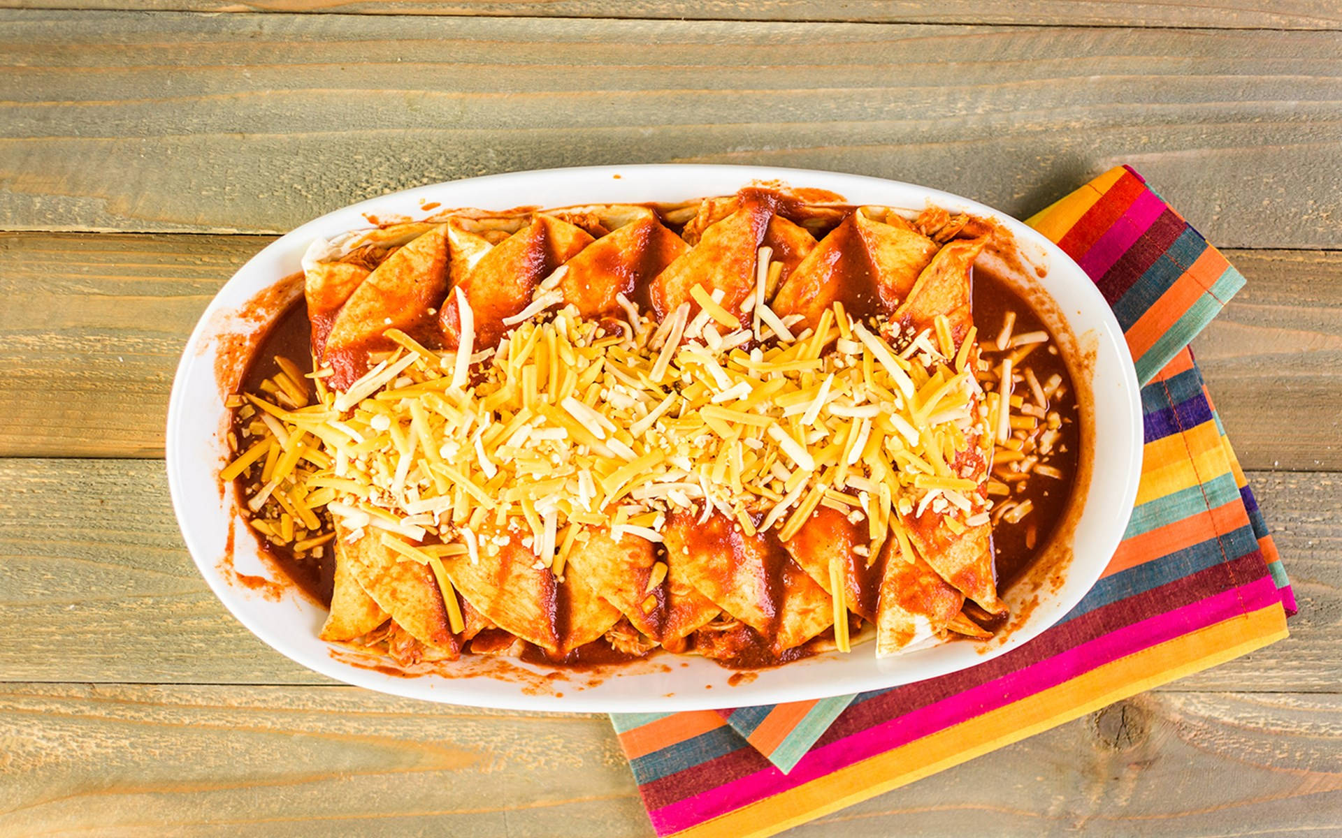 配切達起司的美味 enchiladas 盤 壁紙 — 一個白色橢圓形碟子盛載著金黃色粟米片，上面淋上紅醬和撒上碎芝士，放在一張質樸的木桌上。