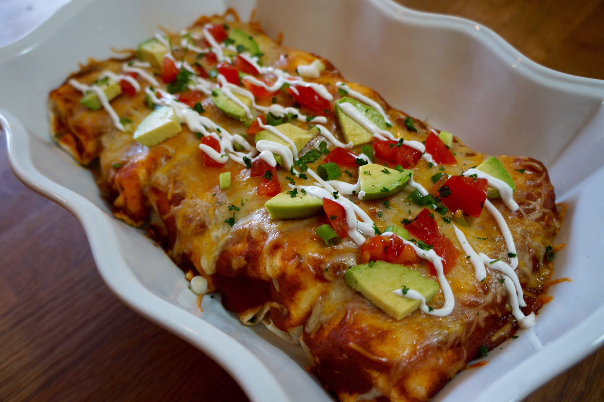 配新鮮牛油果與番茄的美味 enchiladas 壁紙 — 金黃色的玉米捲餅，淋上融化芝士，配上新鮮牛油果、番茄和芫荽，盛載於白色陶瓷碟中。