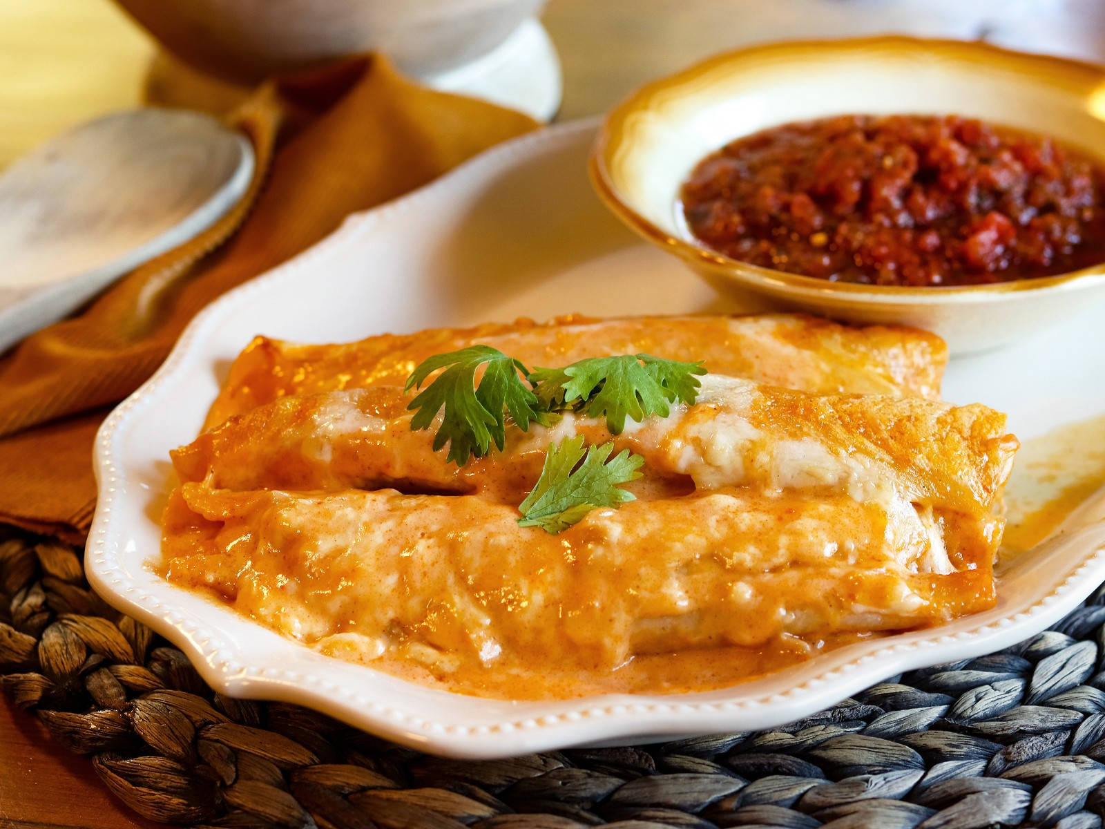 配莎莎醬碗的芝士 enchiladas 盤 壁紙 — 金黃色的玉米捲餅淋上奶油般的紅醬和新鮮香菜，放在白色盤子上，旁邊有一碗粗粒莎莎醬。