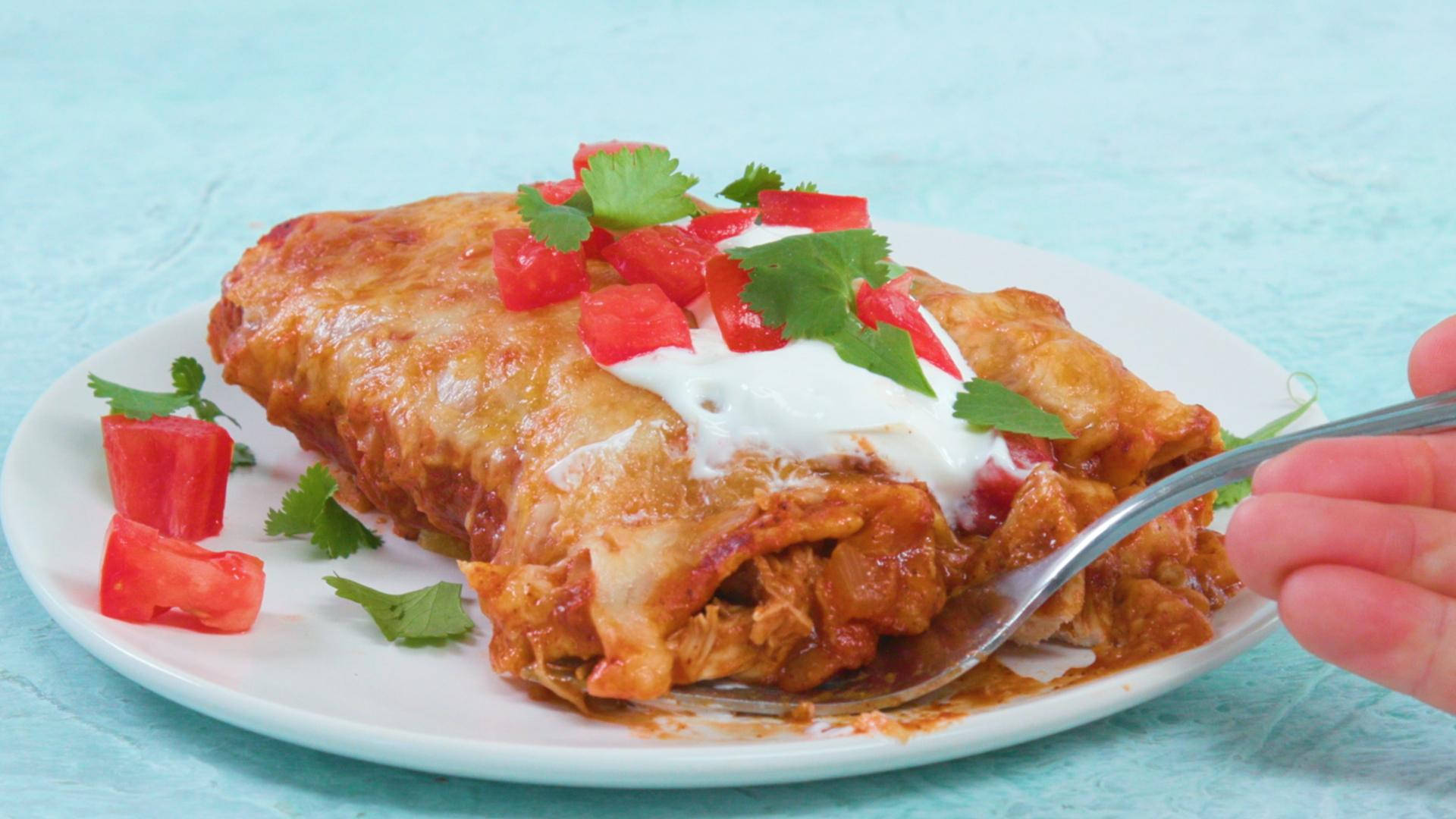 配酸奶油和番茄的 enchiladas 壁紙 — 金黃色雞肉玉米捲餅，上面鋪滿融化芝士、酸忌廉、番茄粒和芫荽，放在白色碟上，有一隻手拿著叉。