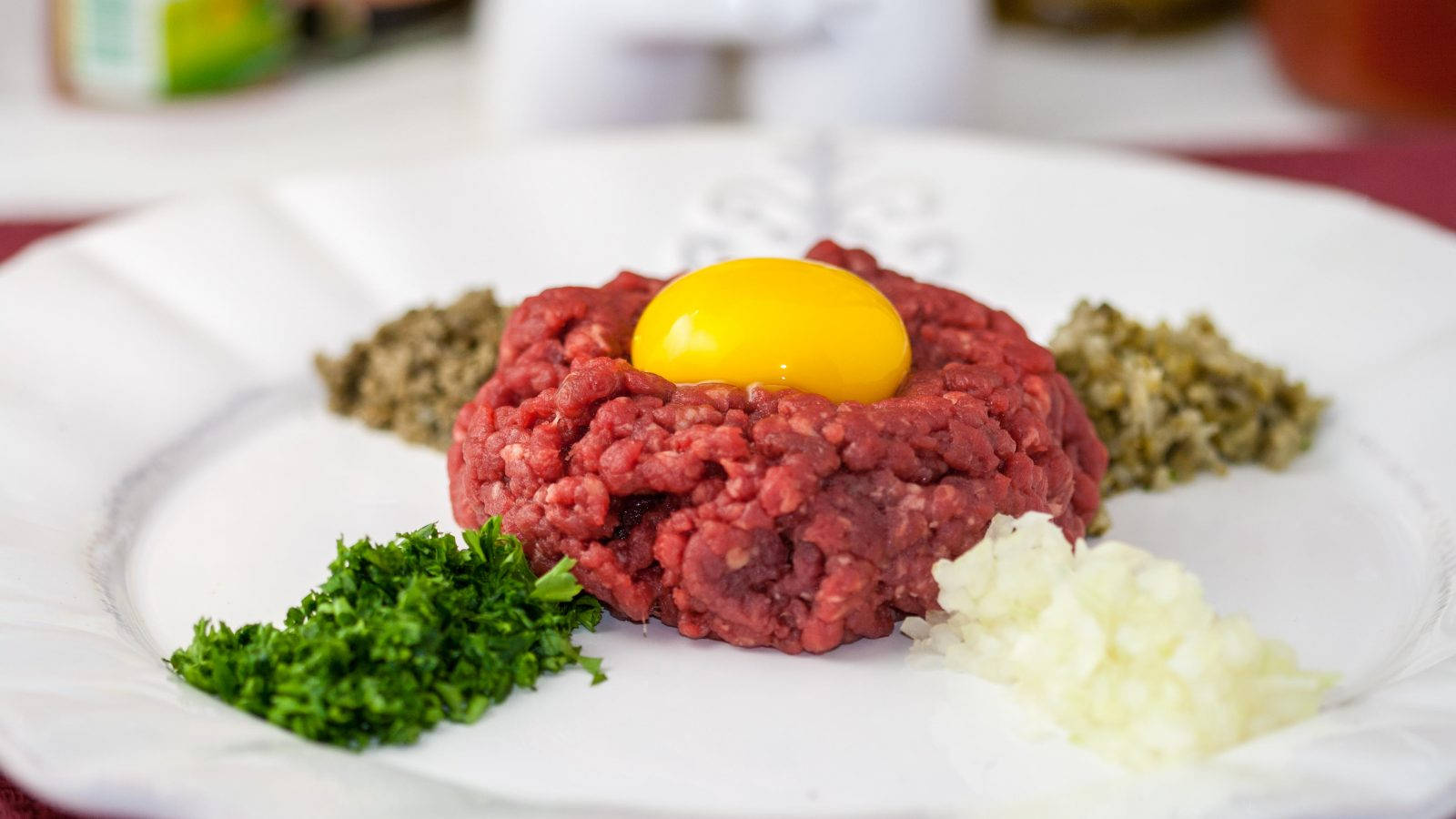 配香草的牛柳塔塔 壁紙 — 一個白色碟子盛載生牛肉他他，中央放有鮮黃色蛋黃，周圍綴以切碎嘅番茜、酸豆同洋蔥。