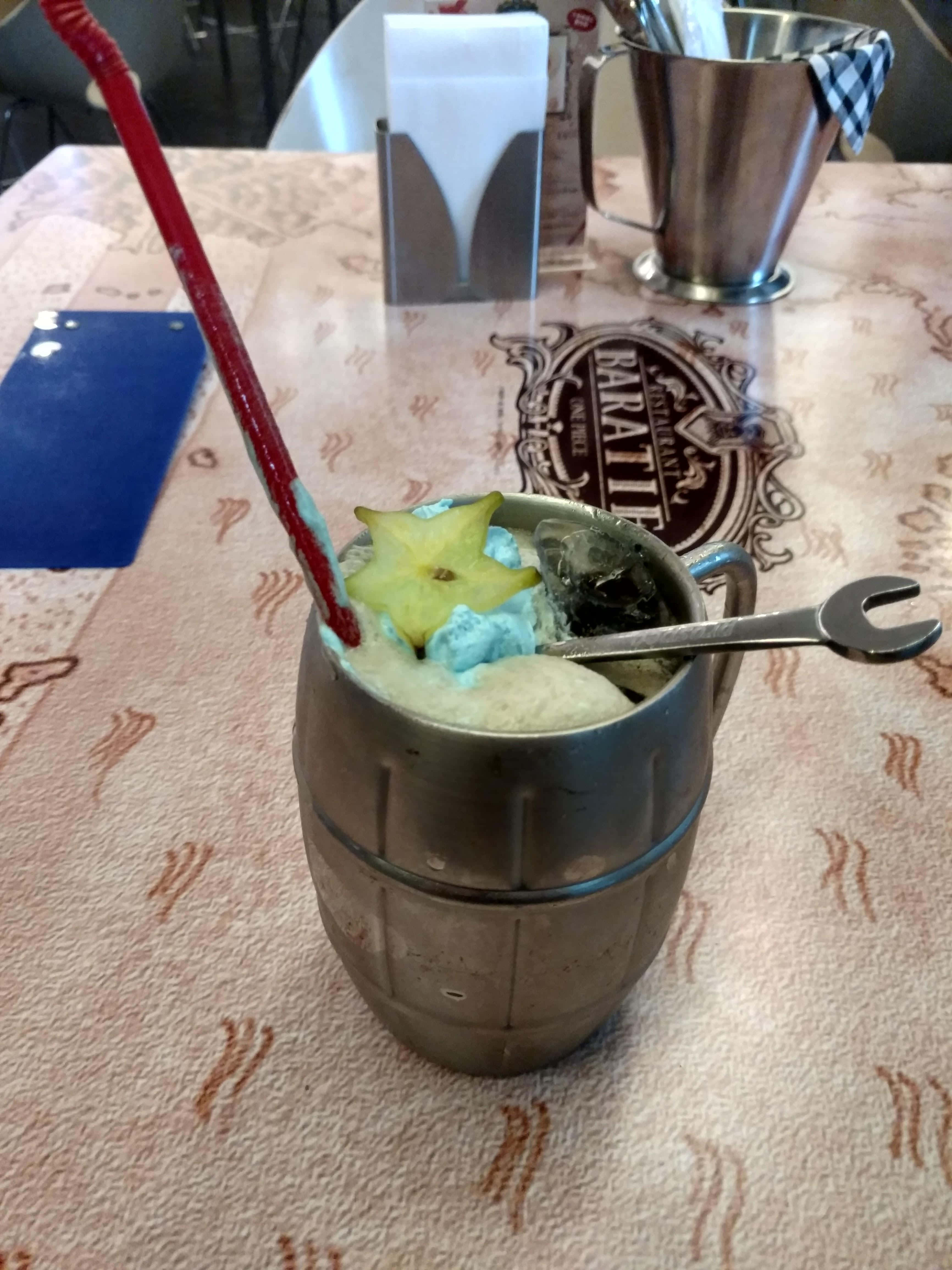 Baratie 桌布 壁紙 — 酒吧內，一個金屬杯盛載著冰凍藍色飲品，上面放有楊桃和紅色飲管，放在圖案枱布上。