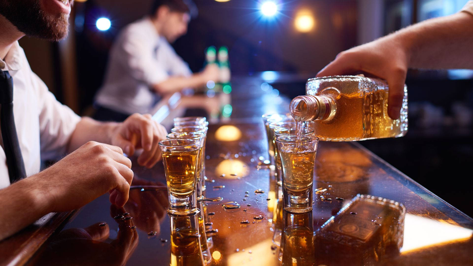 酒吧內燃燒的高級酒品之夜 壁紙 — 調酒師在光亮的吧檯上將琥珀色烈酒倒入小酒杯，一位留鬍鬚的顧客在昏暗的夜總會中觀看。
