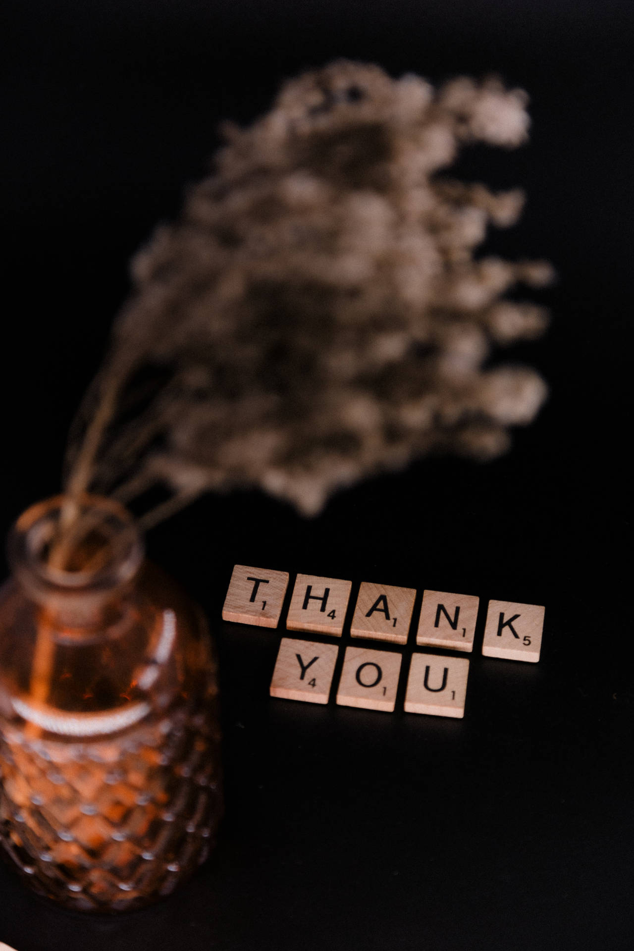 拼出 thank you 的 Scrabble 字母塊放在一個裝飾性琥珀色花瓶旁邊，花瓶裡有乾花。
