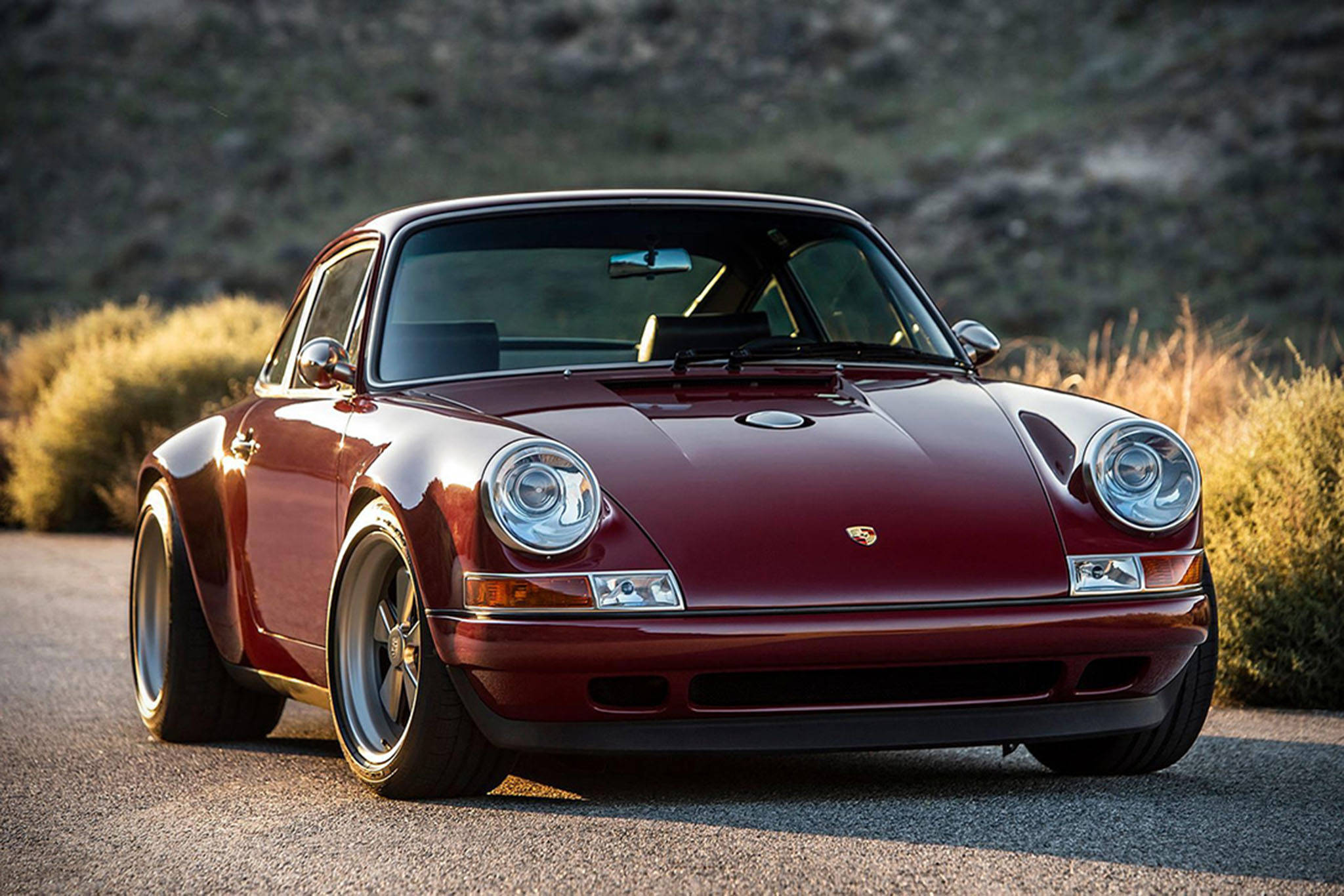 酒紅Singer Porsche 壁紙 — 一架光澤的栗色保時捷911經典跑車停泊在碎石路上，旁邊有乾燥灌木，沐浴在溫暖的金色陽光下。