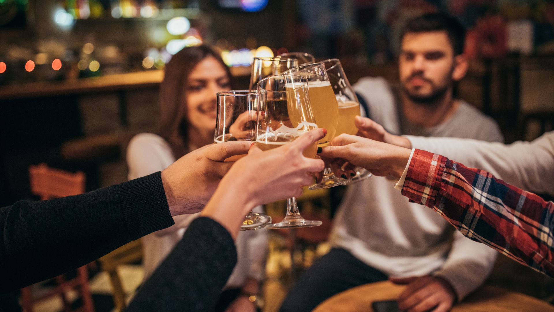 酒館朋友慶祝乾杯攝影 壁紙 — 朋友們在溫暖燈光的酒吧中碰啤酒杯，背景燈光模糊，臉帶微笑。