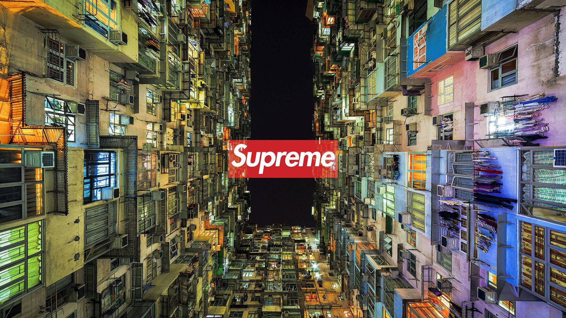 酷 Supreme 建築中庭 壁紙 — 夜晚，狹窄的小巷兩旁是高聳密集的城市建築，上方虛空中置有紅色Supreme標誌。