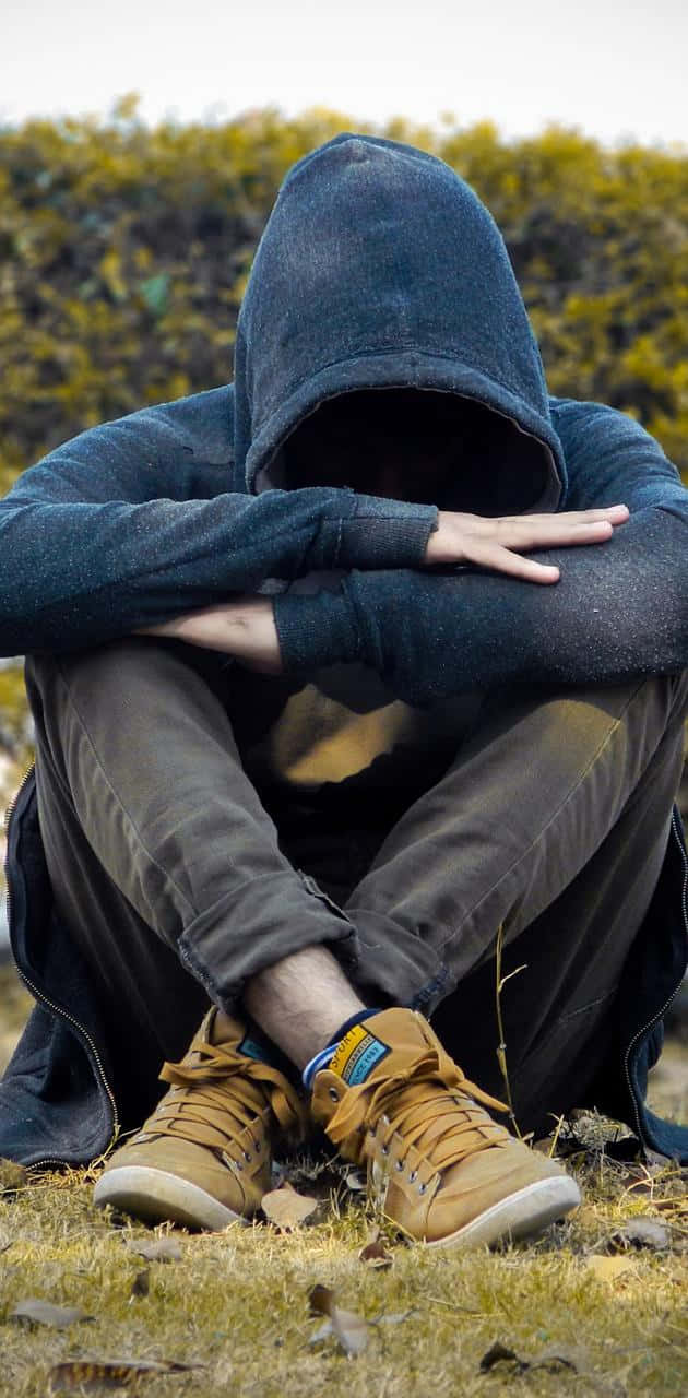 酷帥憂鬱男孩坐在草地 壁紙 — 一個穿著深色連帽衫的人蜷縮在草地上，雙臂環抱膝蓋，臉部隱藏，傳達孤獨和內省。
