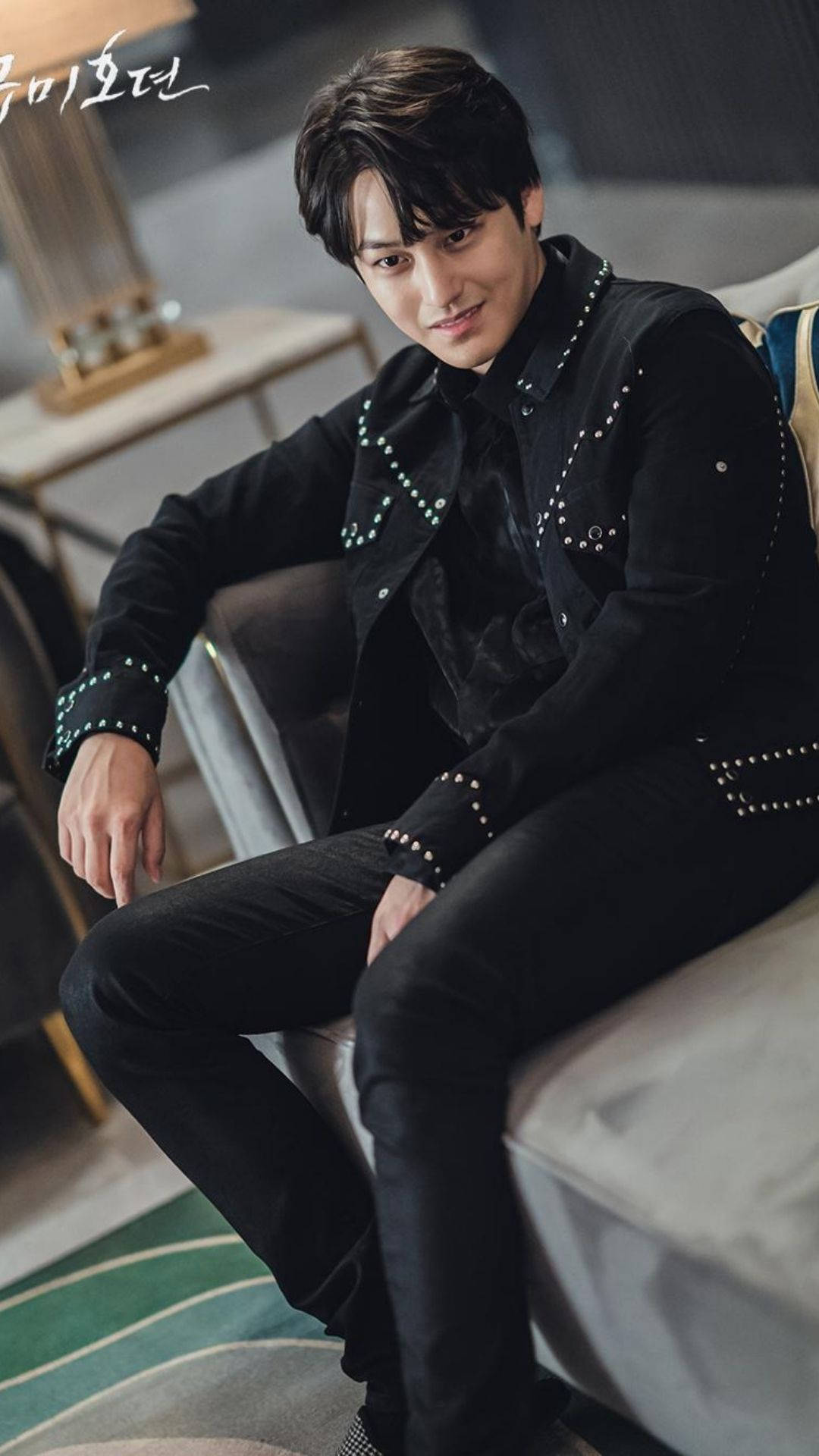 酷帥金汎 壁紙 — 一位時尚男士穿著黑色鉚釘外套，自信地坐在現代沙發上，室內裝潢別緻。
