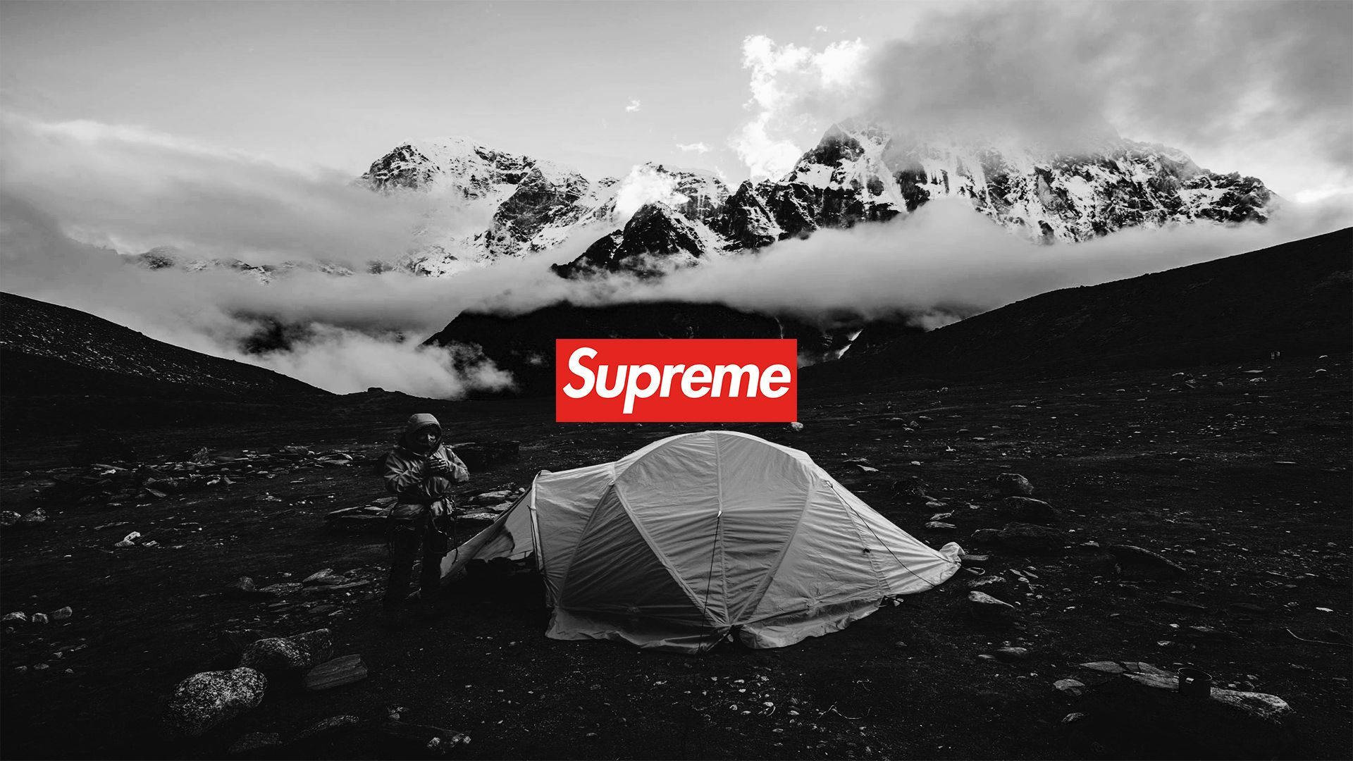 酷炫 Supreme 山間帳篷 壁紙 — 一位孤獨的遠足者在一個荒涼的山谷中搭建帳篷，山谷中有積雪的山峰和雲朵，下方是一個大膽的紅色Supreme標誌。